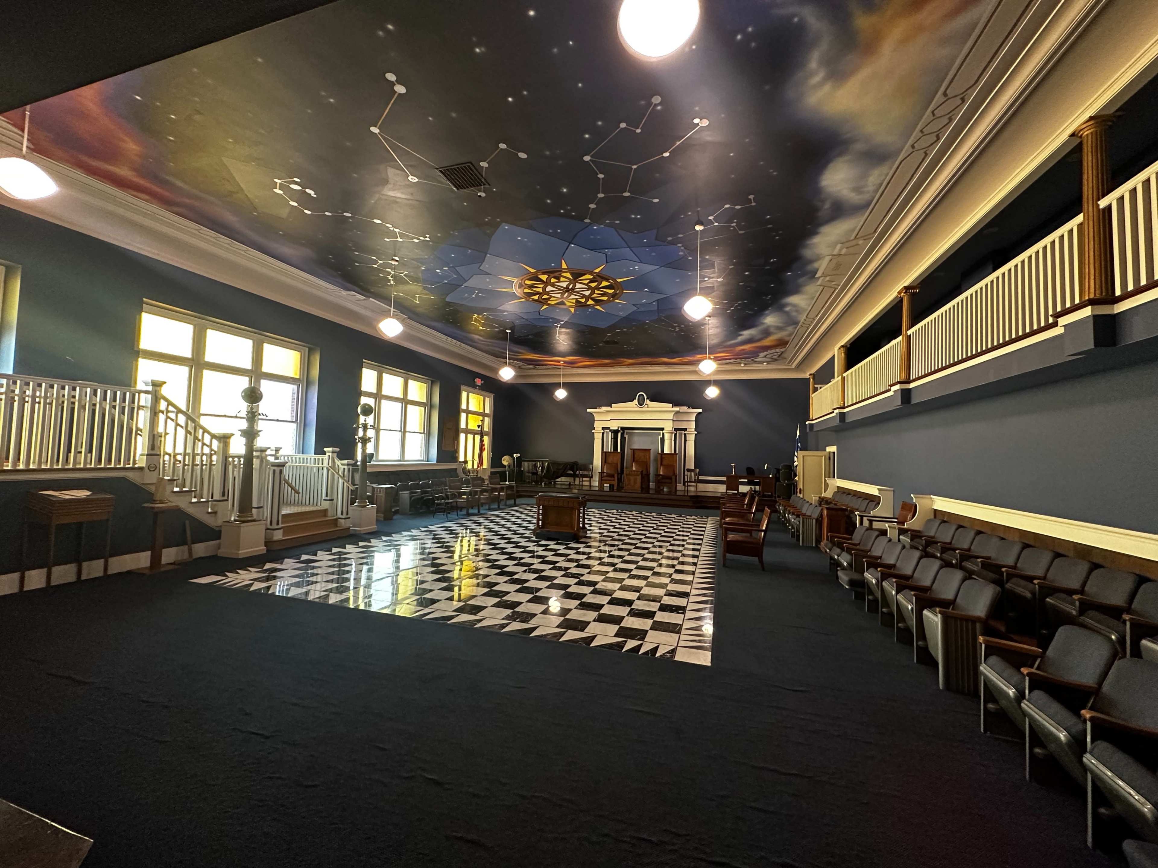 A spacious chamber with a checkered floor, adorned with a celestial-themed mural on the ceiling and rows of wooden seats along the walls.