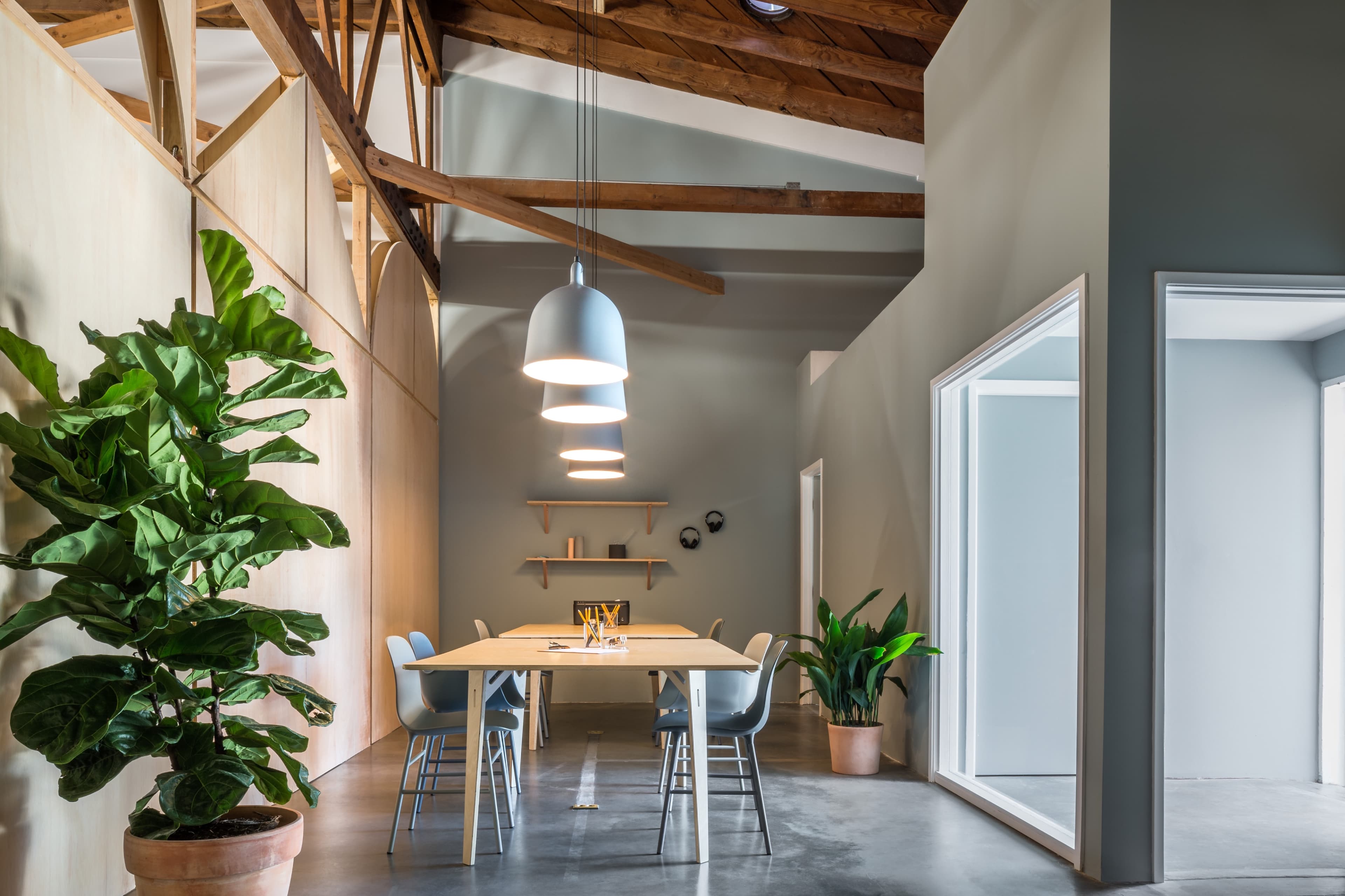 A modern workspace features a table surrounded by chairs, complemented by indoor plants and hanging light fixtures, against a backdrop of wooden beams and gray walls.