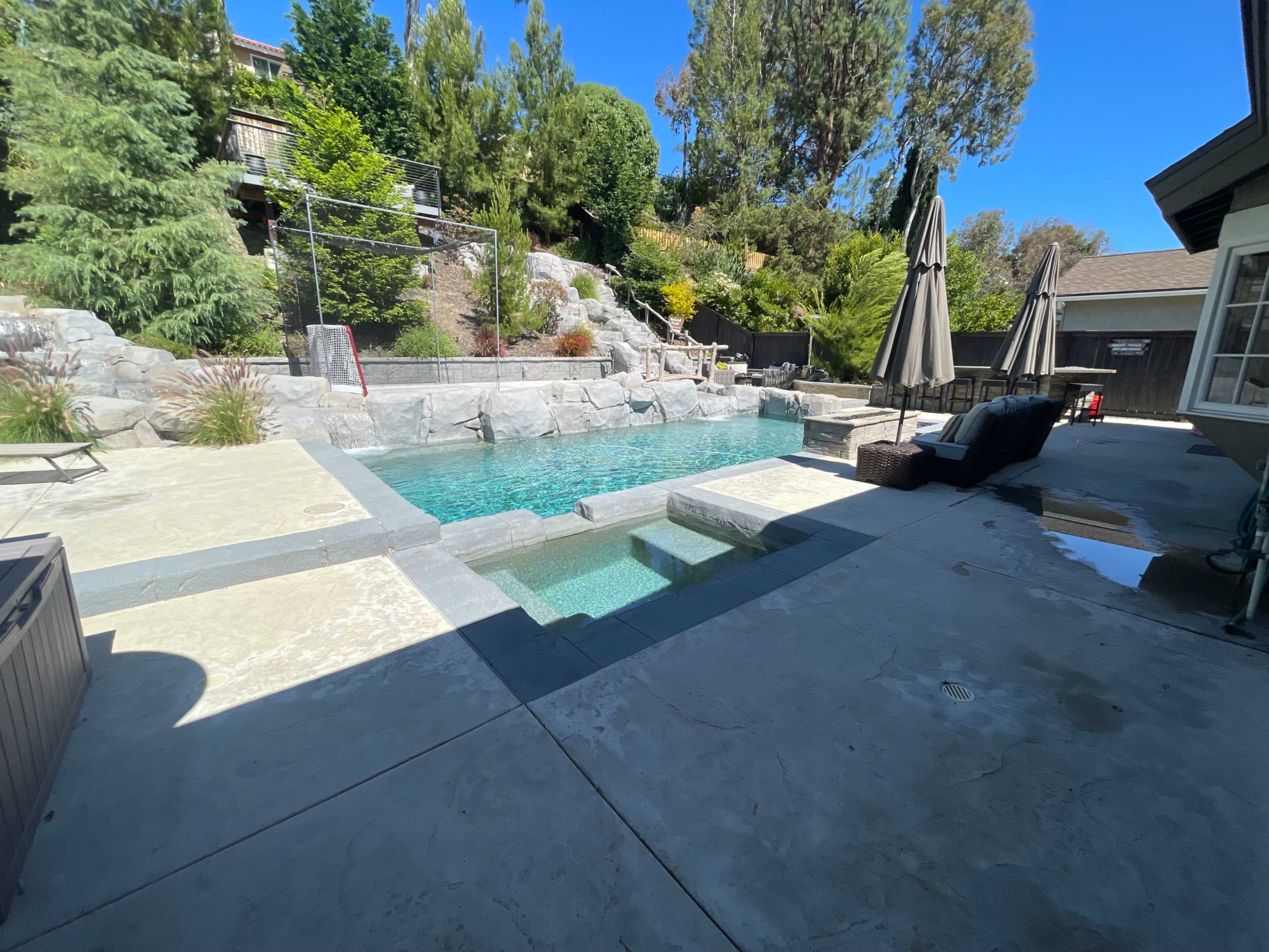 The image shows a swimming pool surrounded by stone landscaping and several patio umbrellas.
