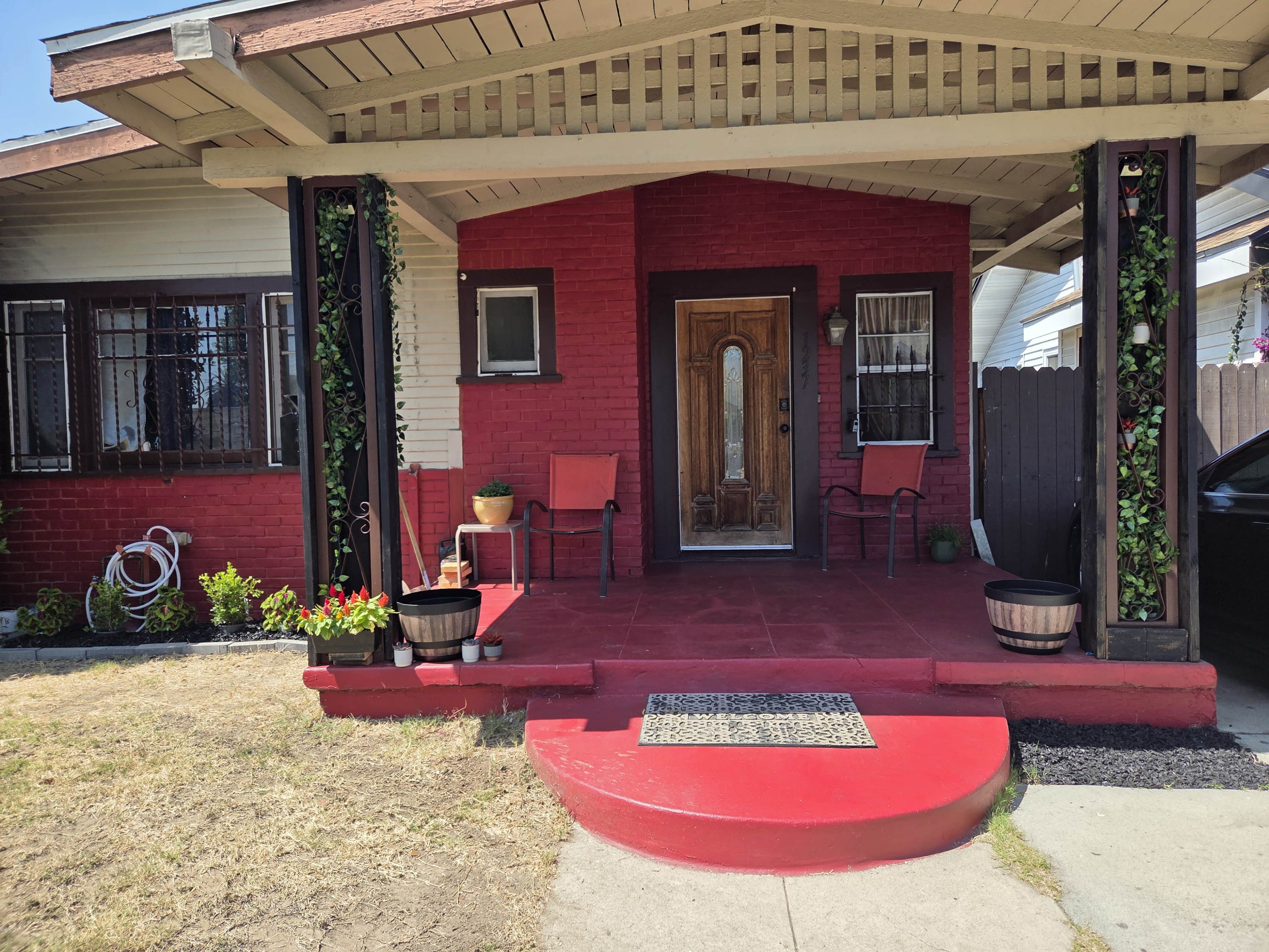The image shows a residential porch with a red accent wall, a wooden front door, two chairs, and potted plants.