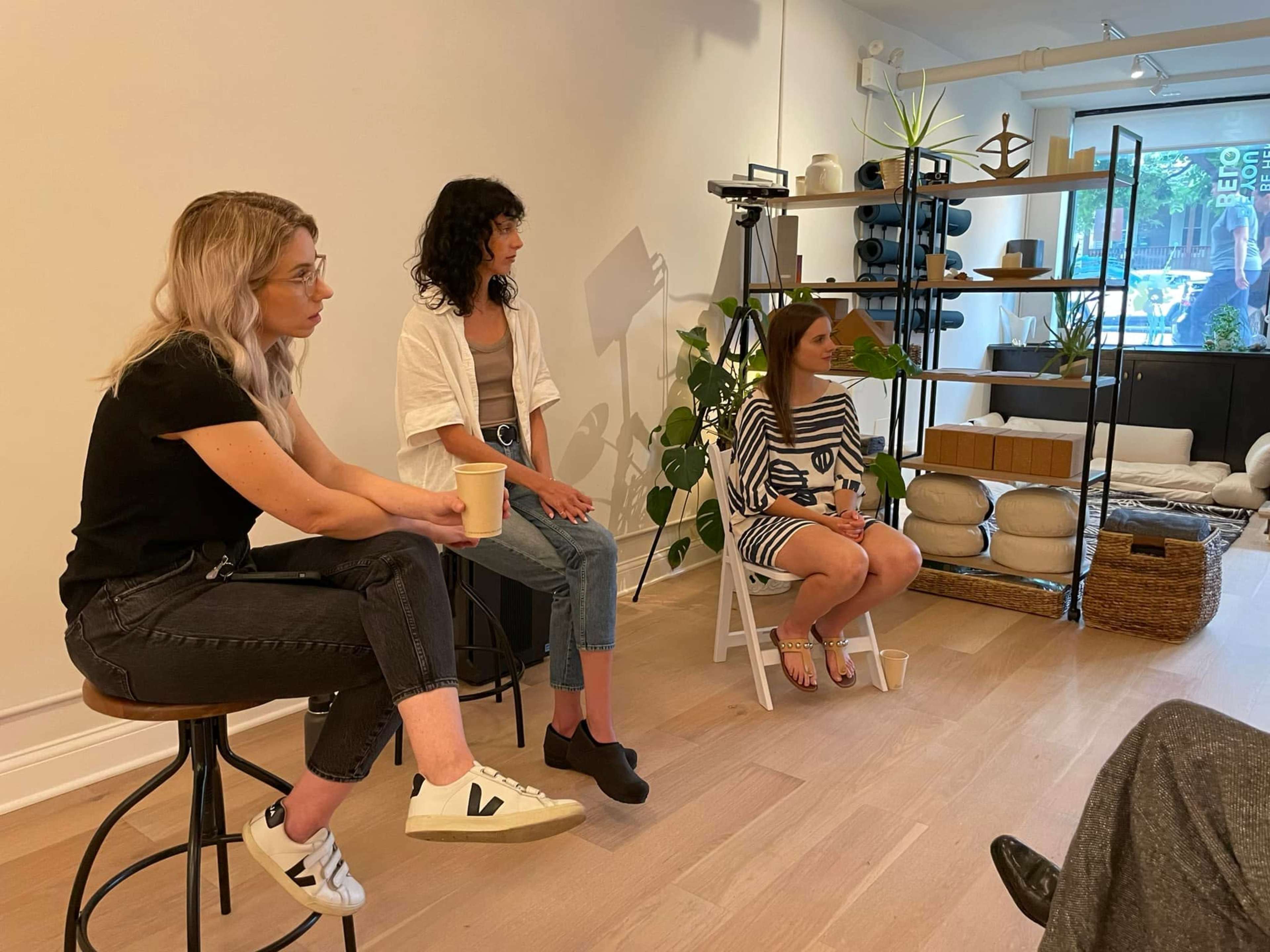 Three women sit on stools and chairs in a minimalistic room, engaged in conversation, with plants and decorative items in the background.
