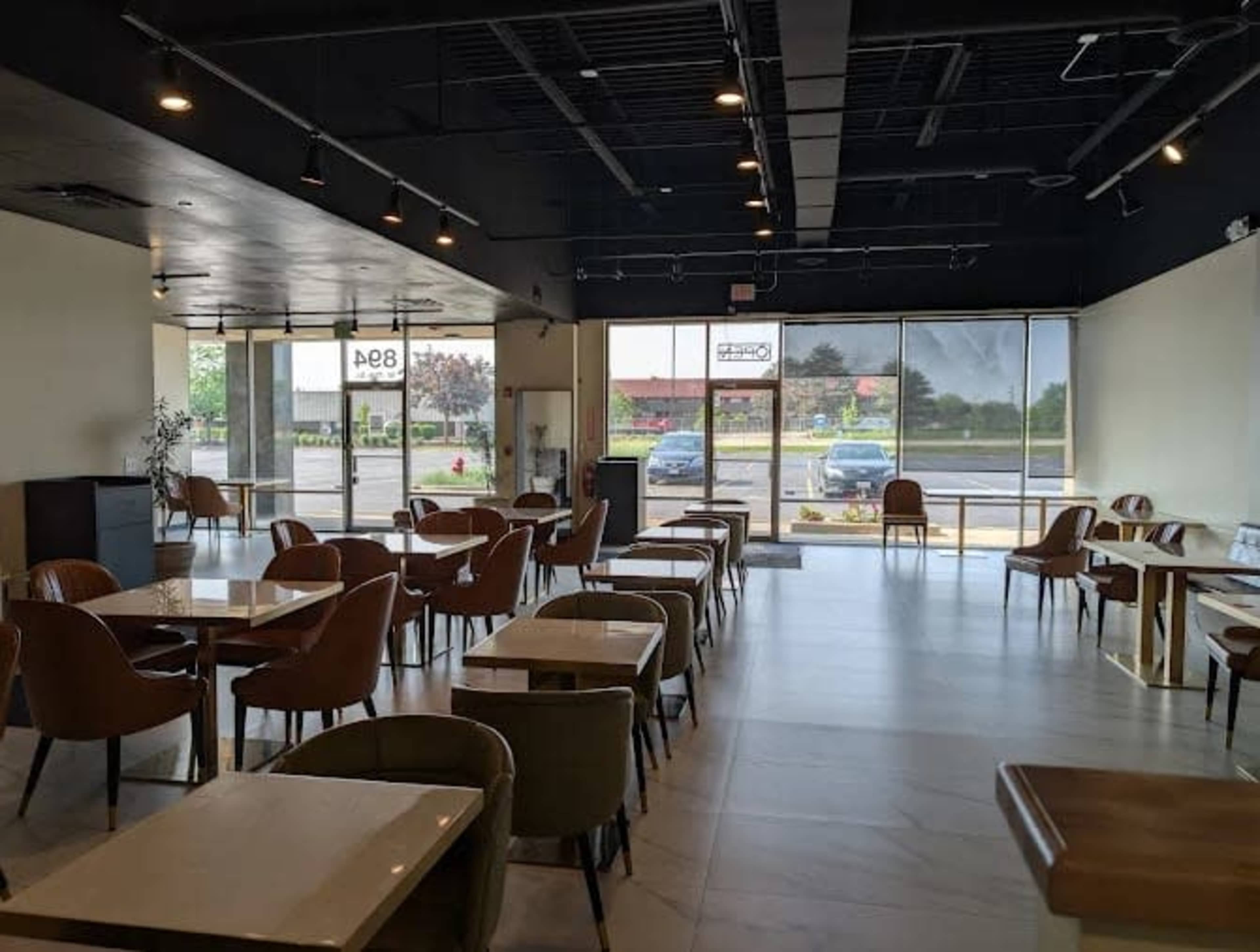 The image shows an interior of a modern café with several tables and chairs, large windows, and a view of the outside.