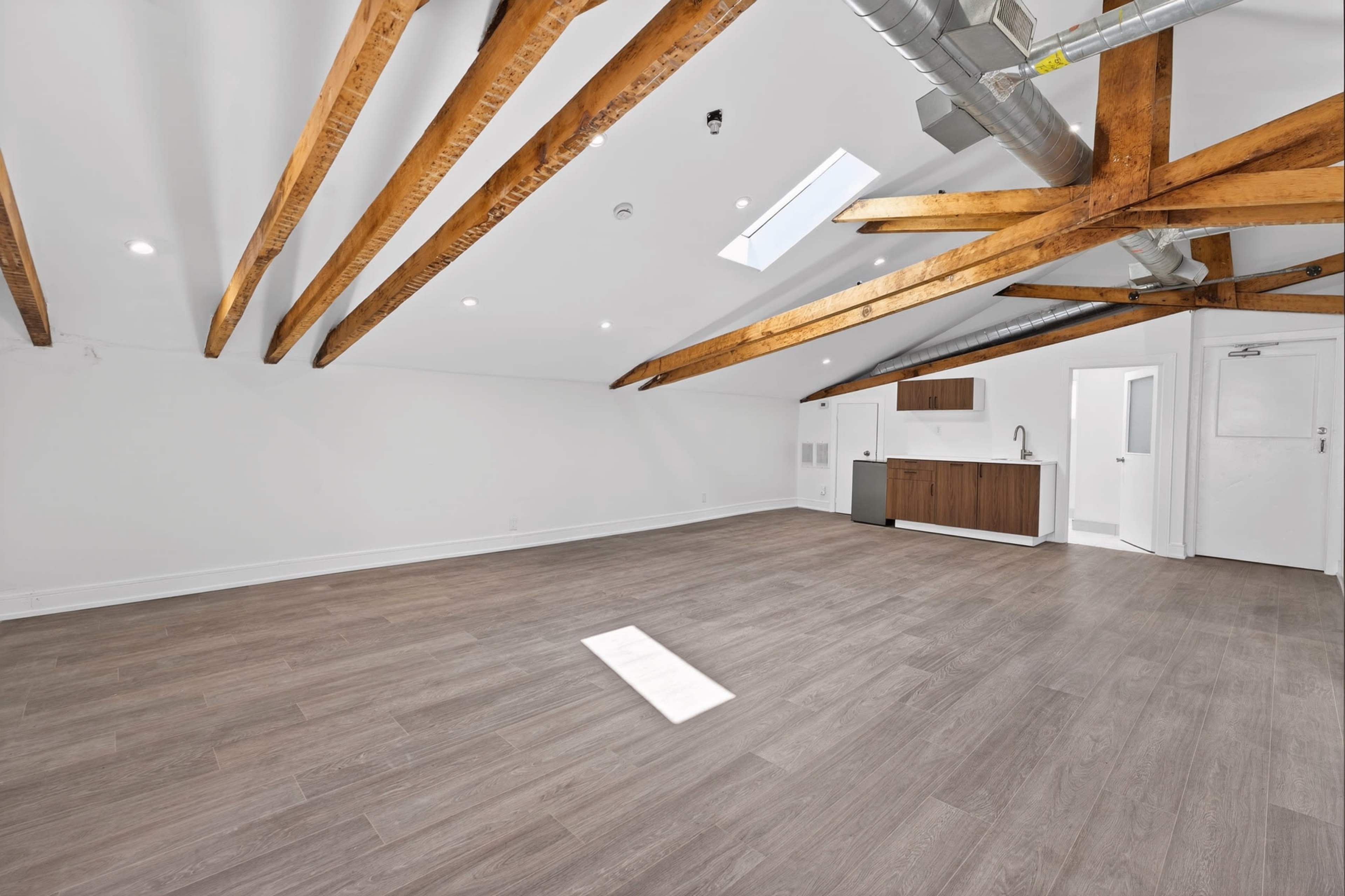 The image shows a spacious room with exposed wooden beams, a skylight, and a kitchenette along one wall.
