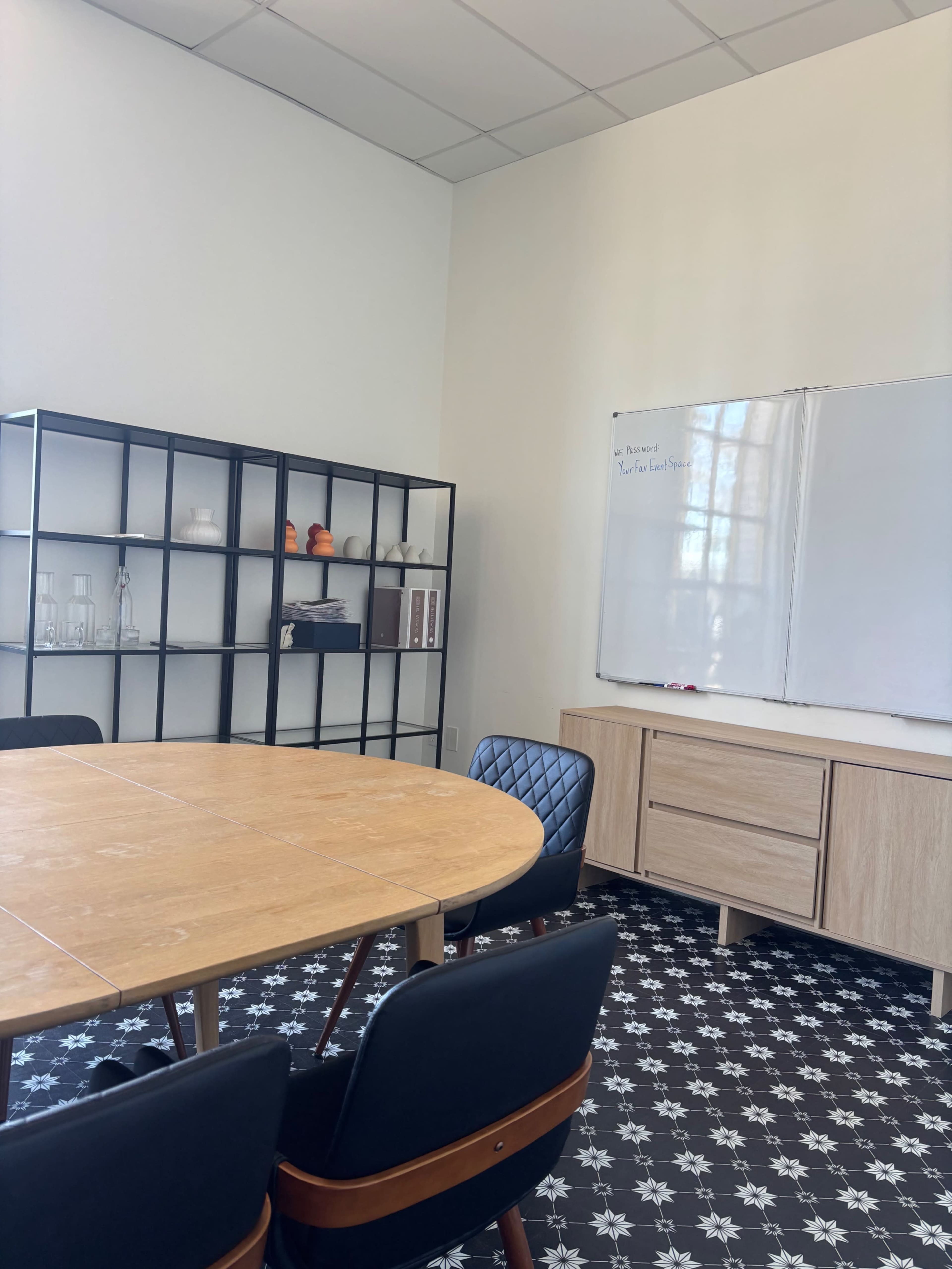 A modern meeting room features a round wooden table surrounded by black chairs, with a whiteboard and shelves displaying glassware and decor.