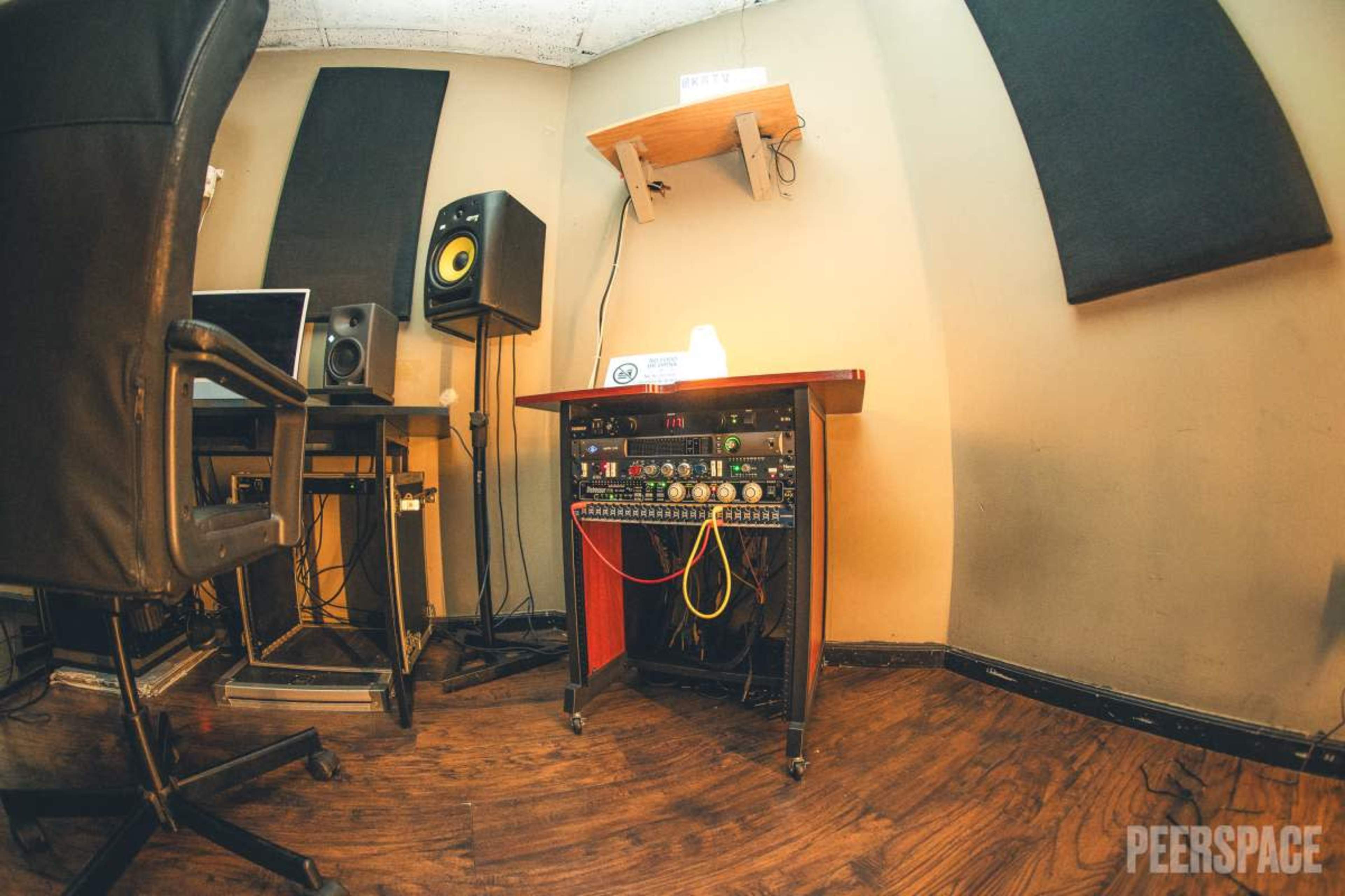 The image depicts a recording studio corner featuring an audio mixer, a computer setup, and soundproofing panels on the walls.
