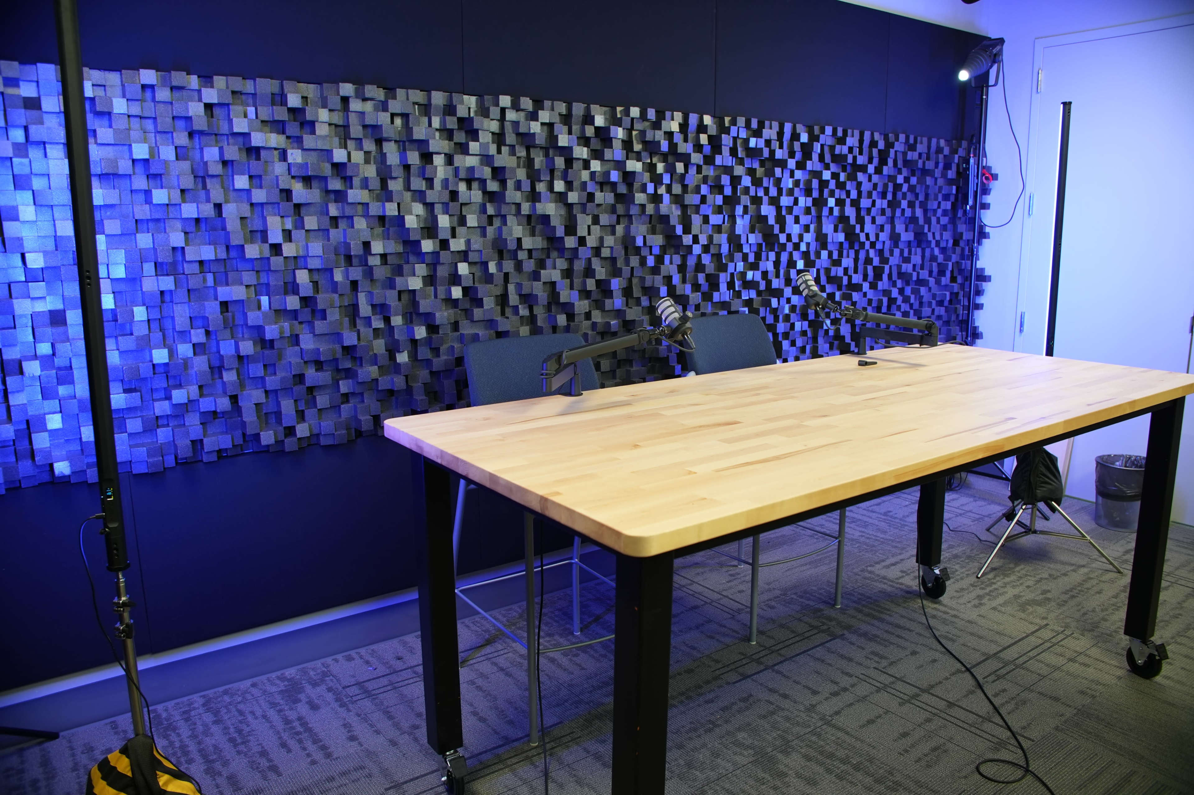 A large wooden table is positioned in front of a textured wall, with two microphones set up on it and blue lighting illuminating the space.
