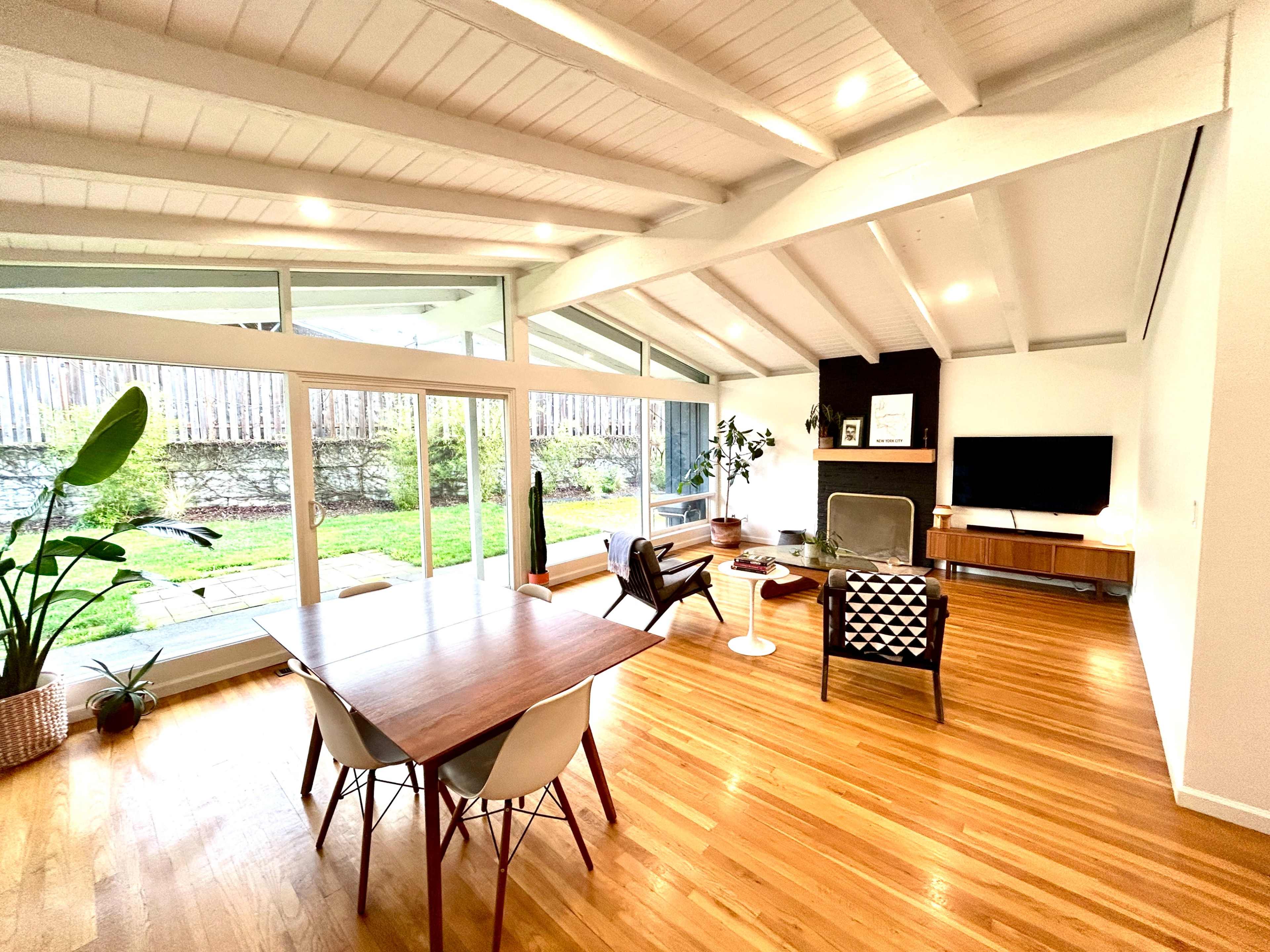 A modern living space features a dining table, a seating area, and large windows overlooking a garden.