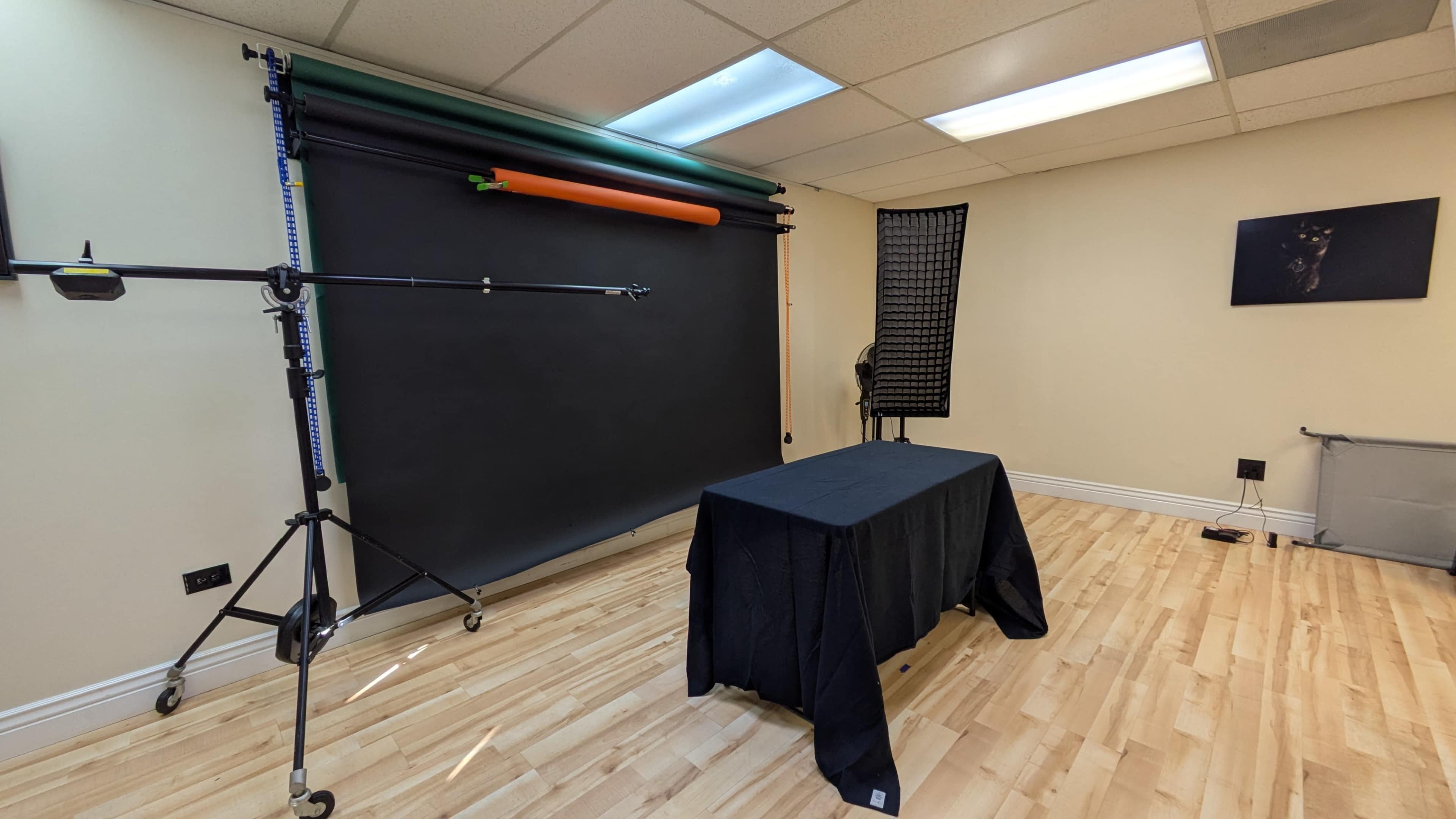 The image shows a photography studio featuring a black backdrop on a stand, a table covered with a black cloth, and a lighting setup in a well-lit room with wooden flooring.