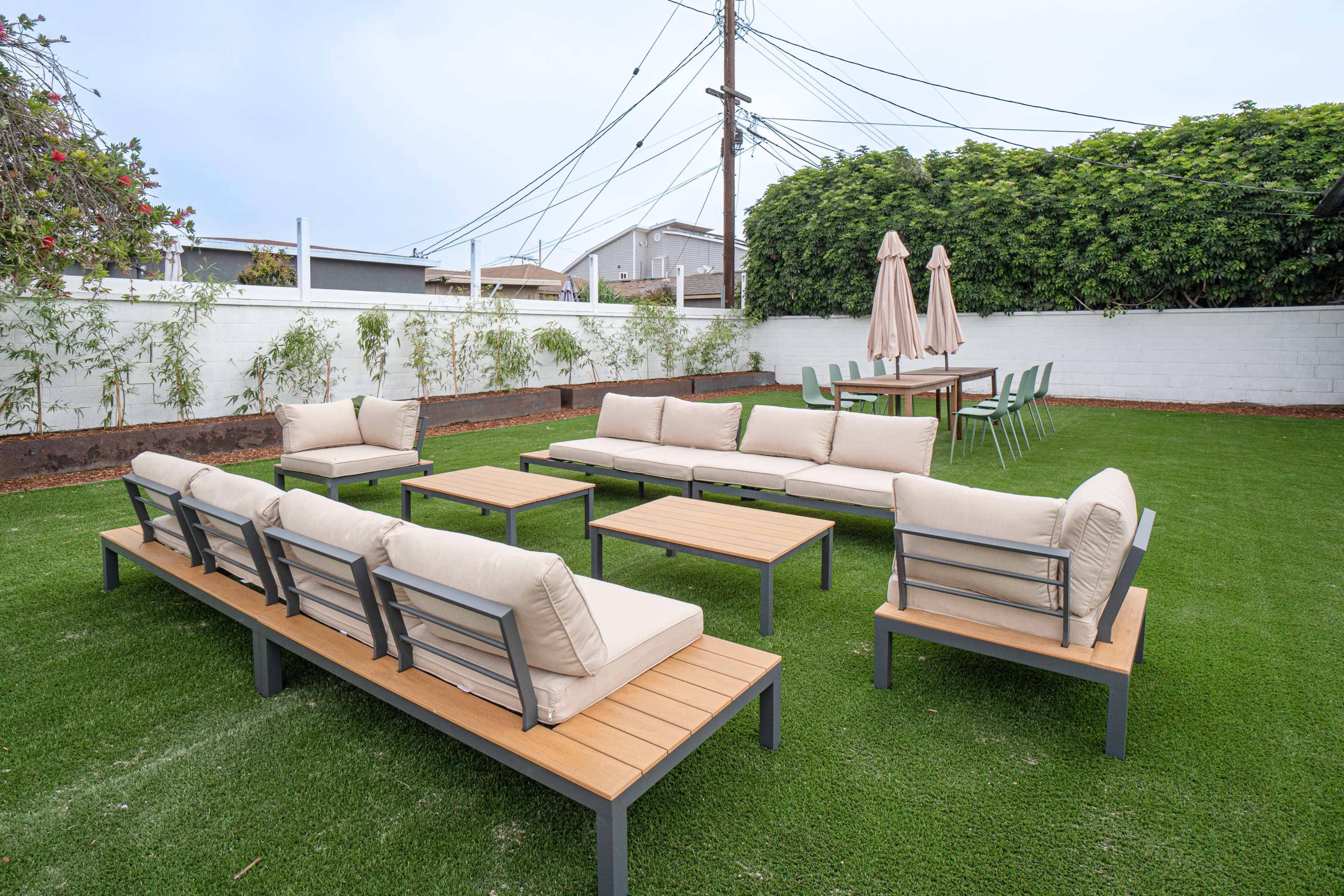 The image shows a landscaped backyard with a seating area featuring two rows of sectional sofas, a coffee table, and a dining table surrounded by chairs, all situated on green grass with a wall and power lines in the background.
