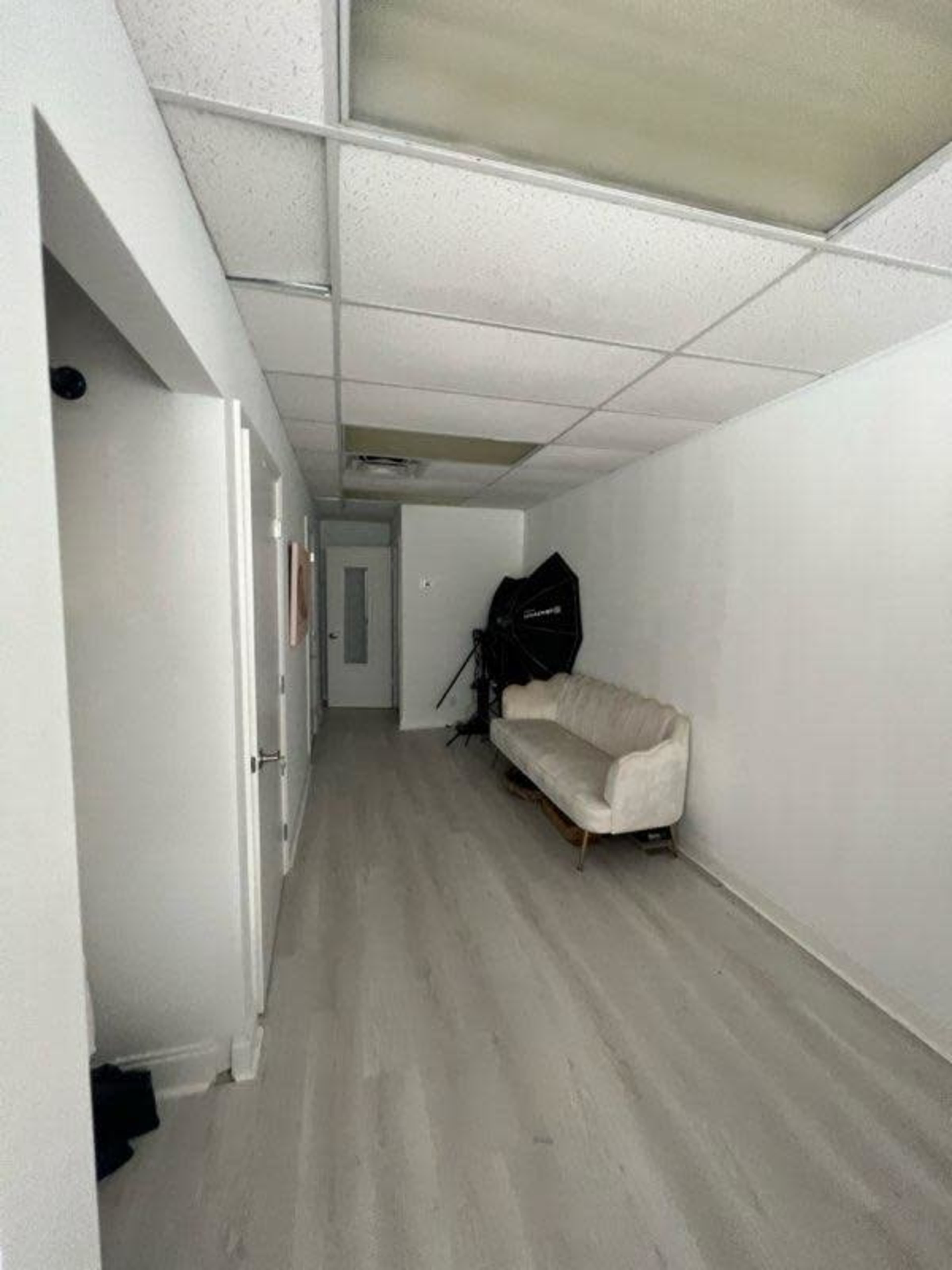 The image shows a narrow hallway with light-colored flooring, a white couch against one wall, and a light fixture on the ceiling.