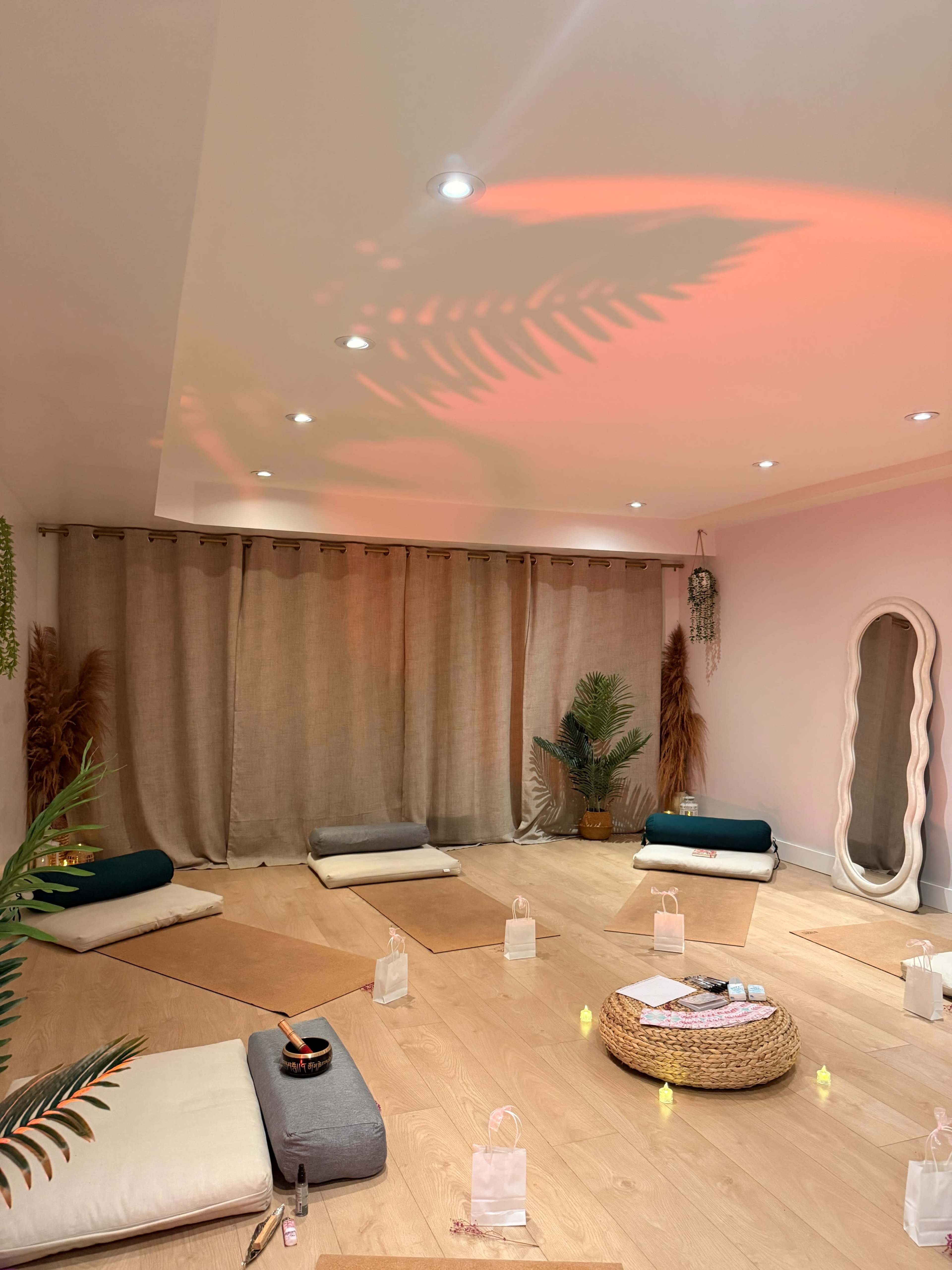 The image shows a calming wellness space with floor cushions arranged around a central basket and soft lighting, accented by green plants and flowing curtains.