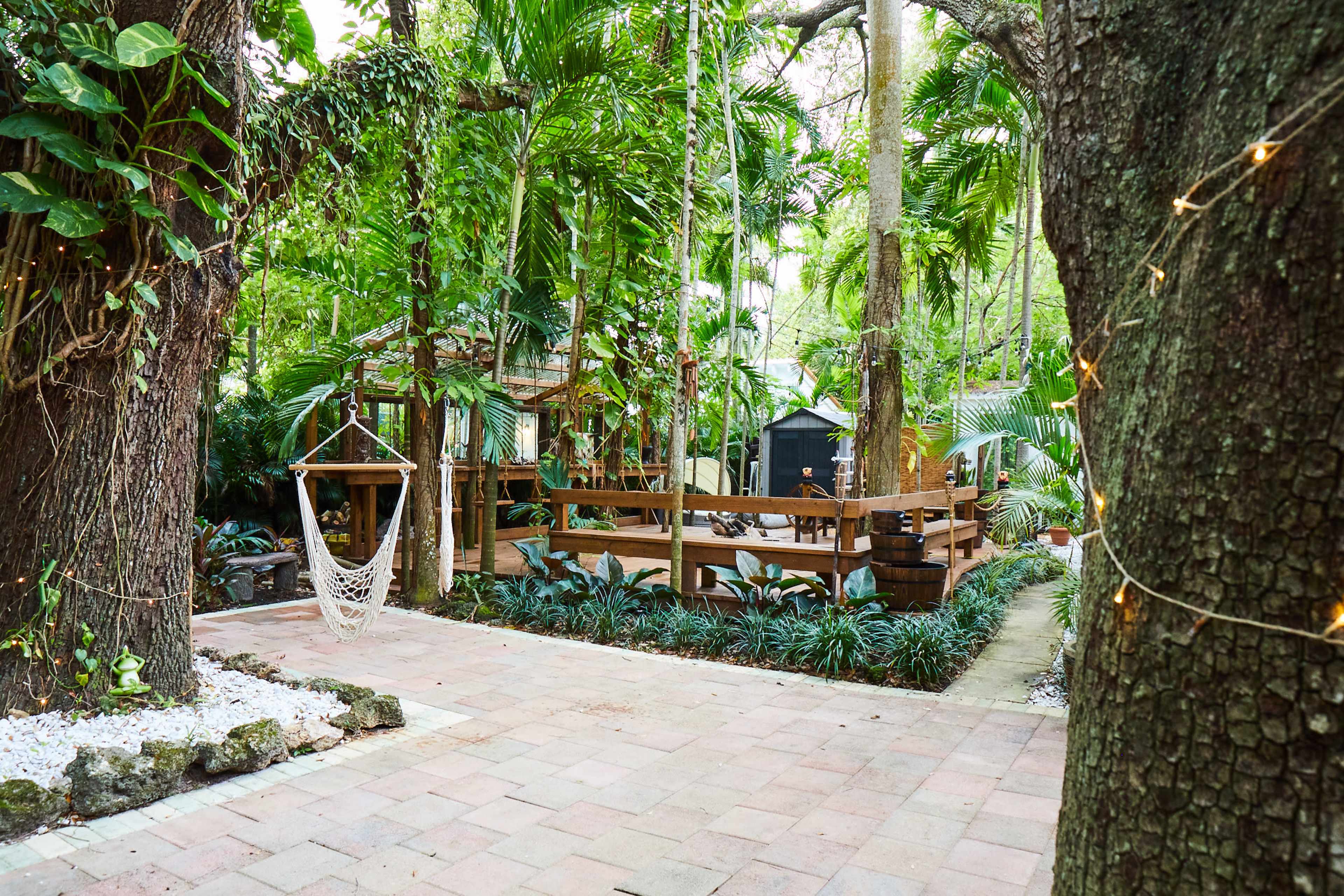 A tropical garden featuring a hammock suspended between two trees, wooden decking, and lush greenery.
