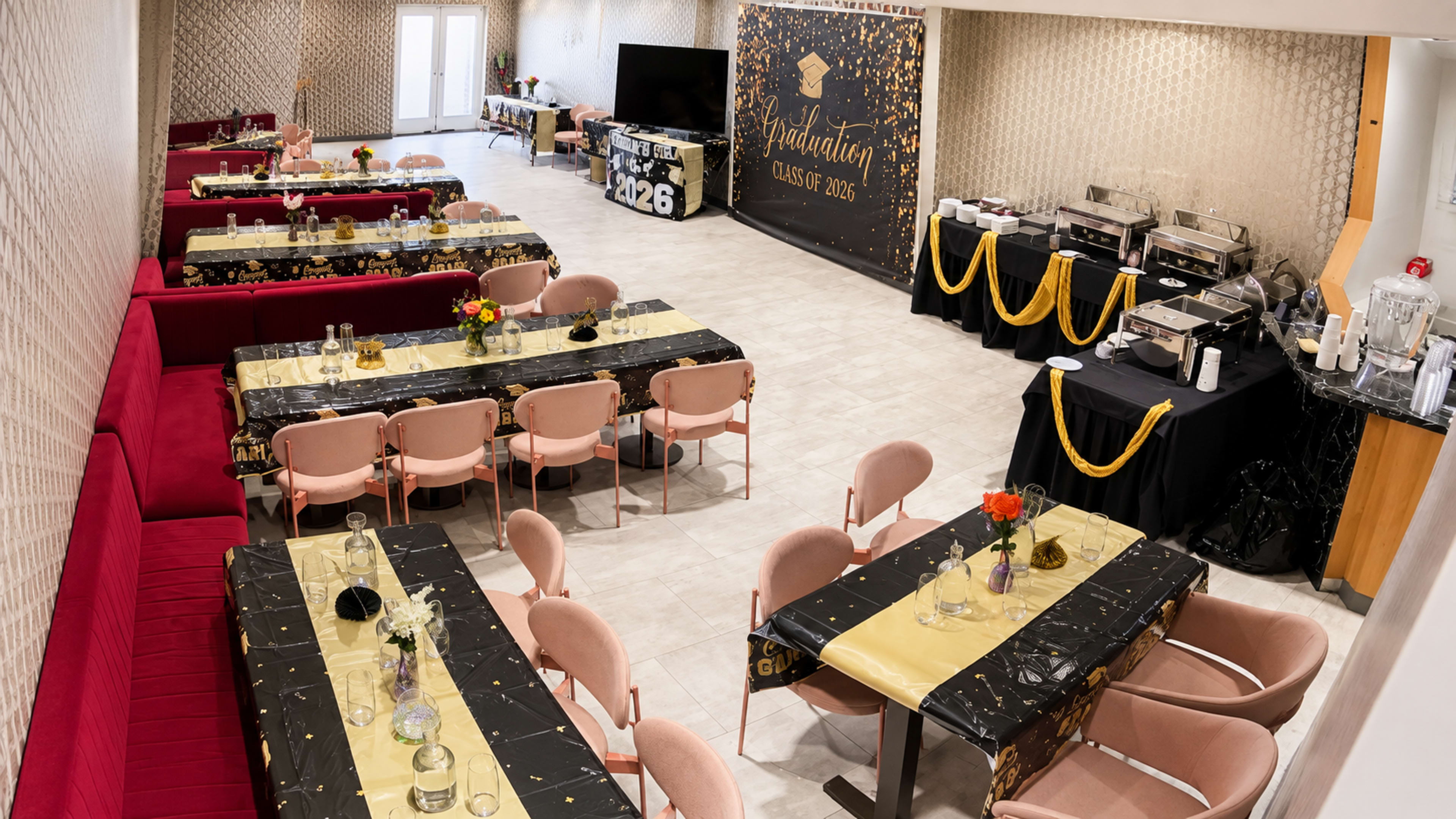 The image shows a decorated banquet hall set up for a graduation celebration, featuring tables with black and gold tablecloths, floral centerpieces, and a backdrop displaying “Graduation Class of 2026.”