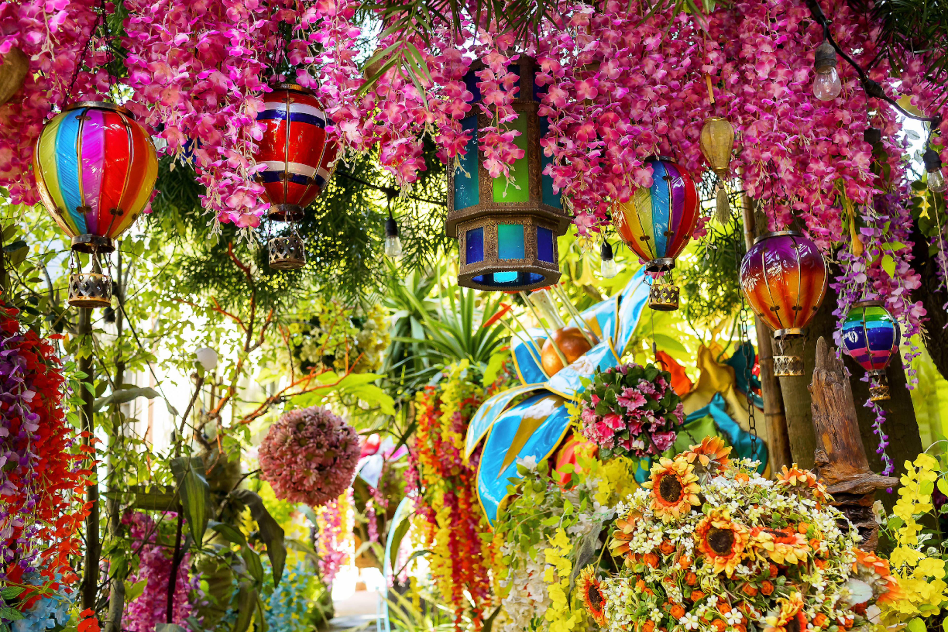 A vibrant scene filled with colorful hanging lanterns and an abundance of artificial flowers in a lush, tropical setting.