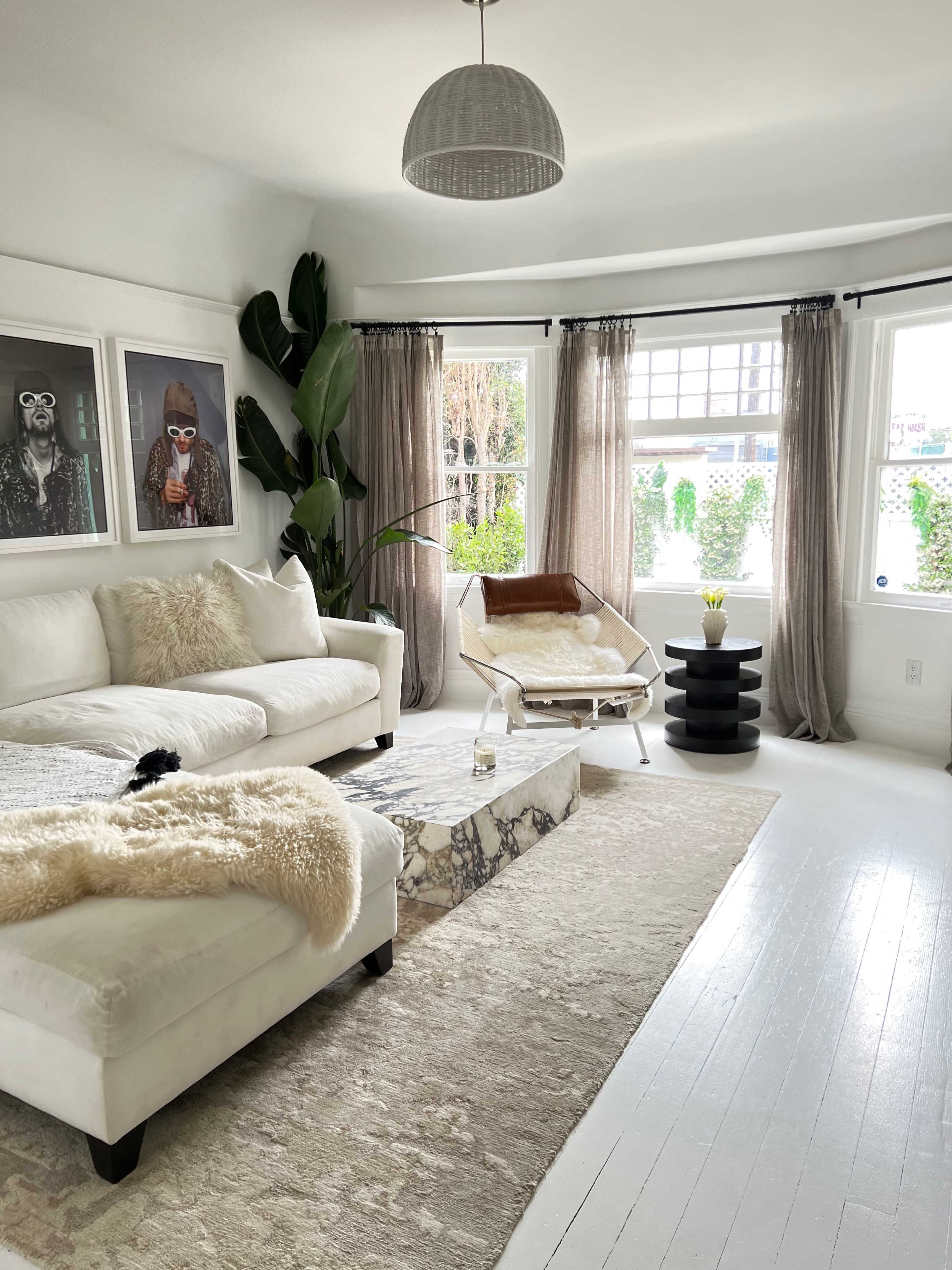 A bright living room features a white sectional sofa, a marble coffee table, and large windows adorned with sheer curtains, showcasing green outdoor plants.