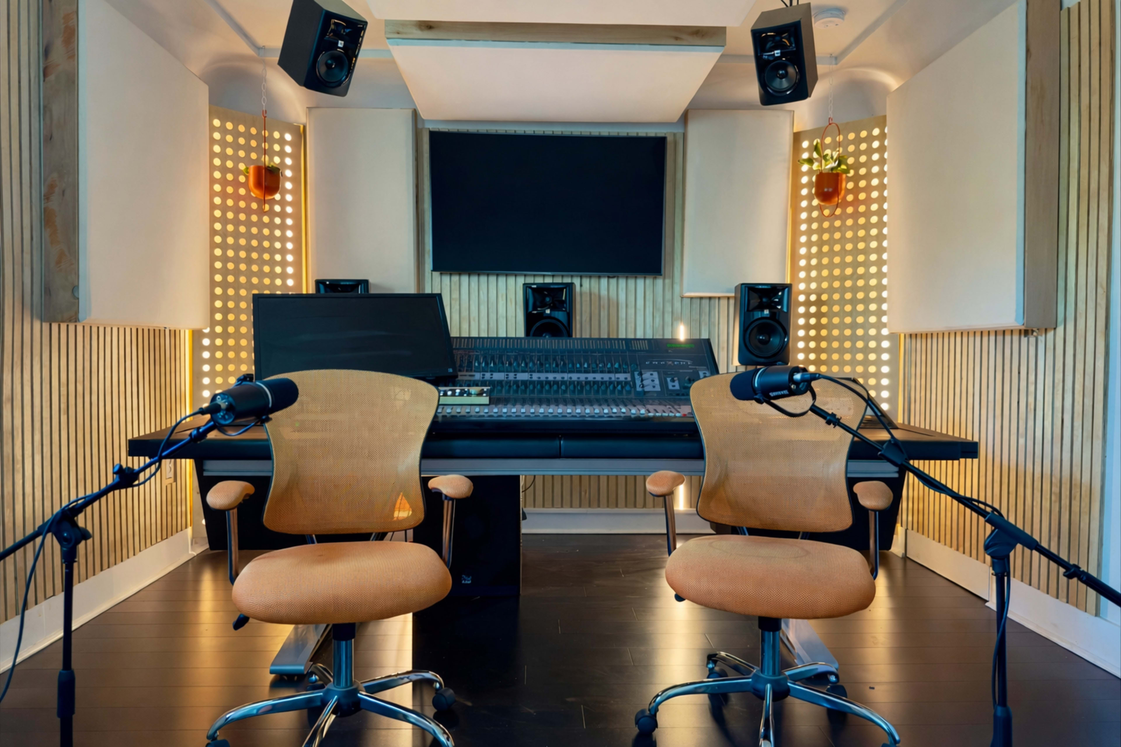 The image shows a modern recording studio with two chairs and a sound mixing console, flanked by studio monitors and acoustic paneling on the walls.