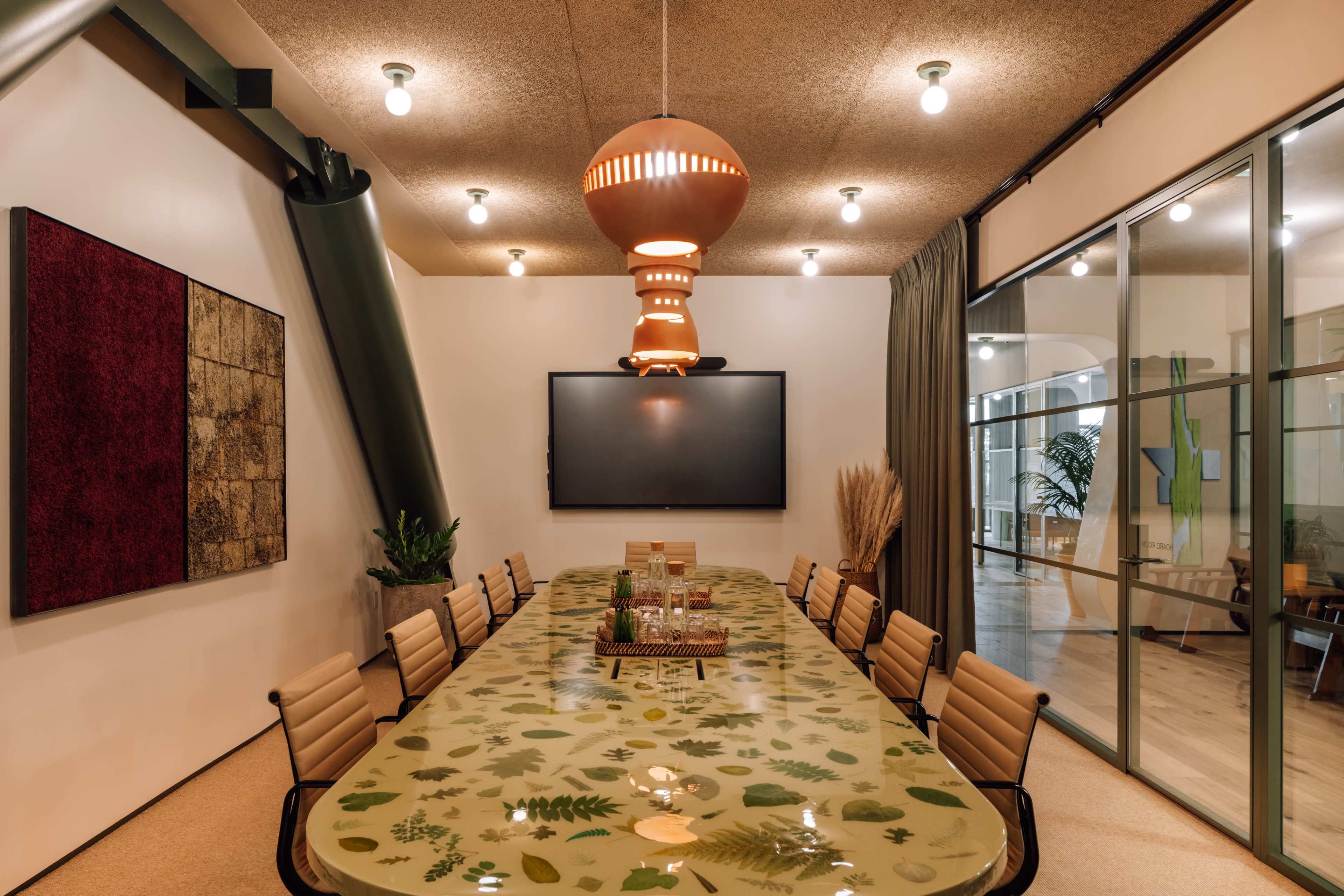 The image depicts a modern conference room featuring a long table with a leaf-patterned surface, surrounded by ergonomic chairs, and equipped with a large screen on one wall.
