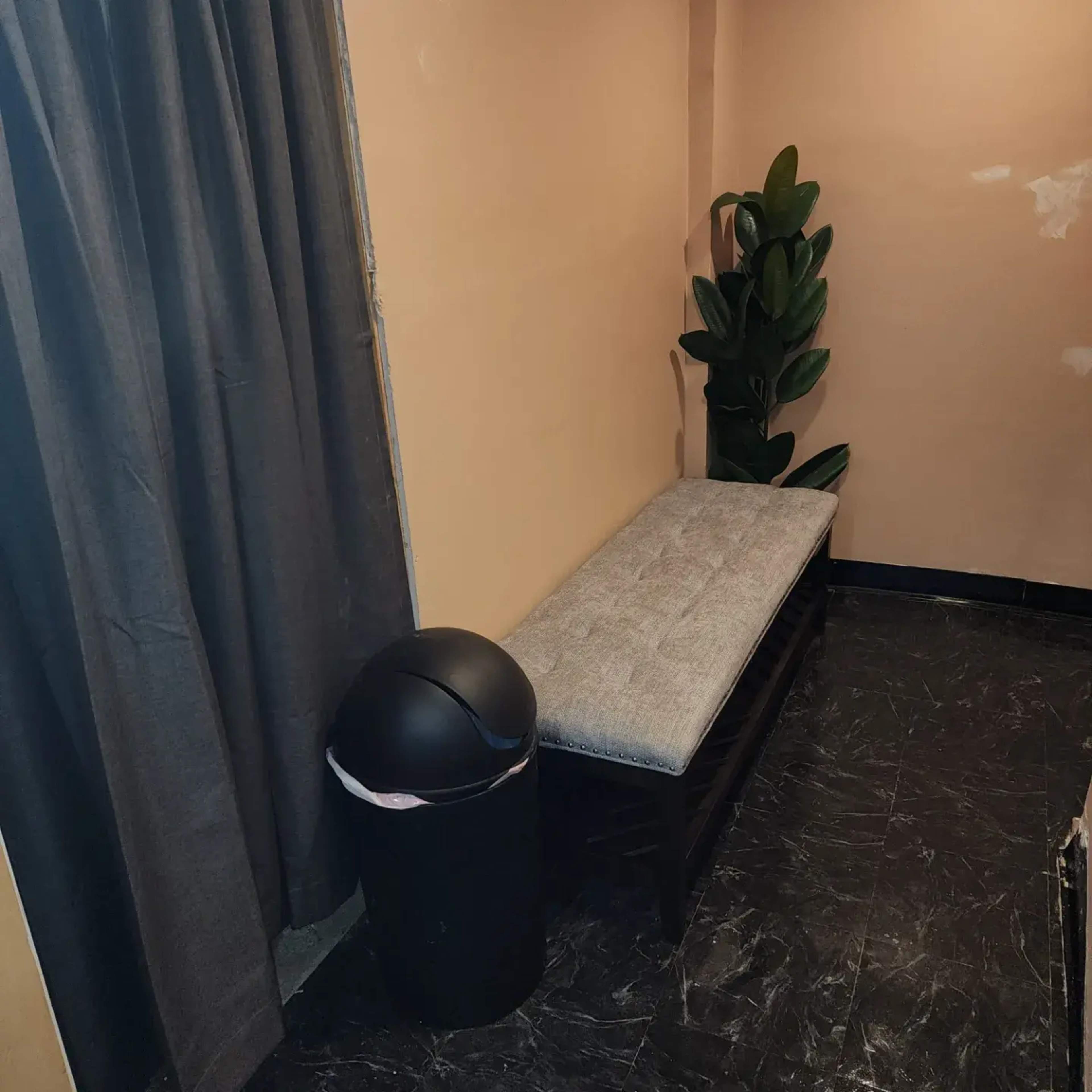 A small corner features a wooden bench with a gray cushion, a black trash can, and a potted plant, against a wall with a curtain.