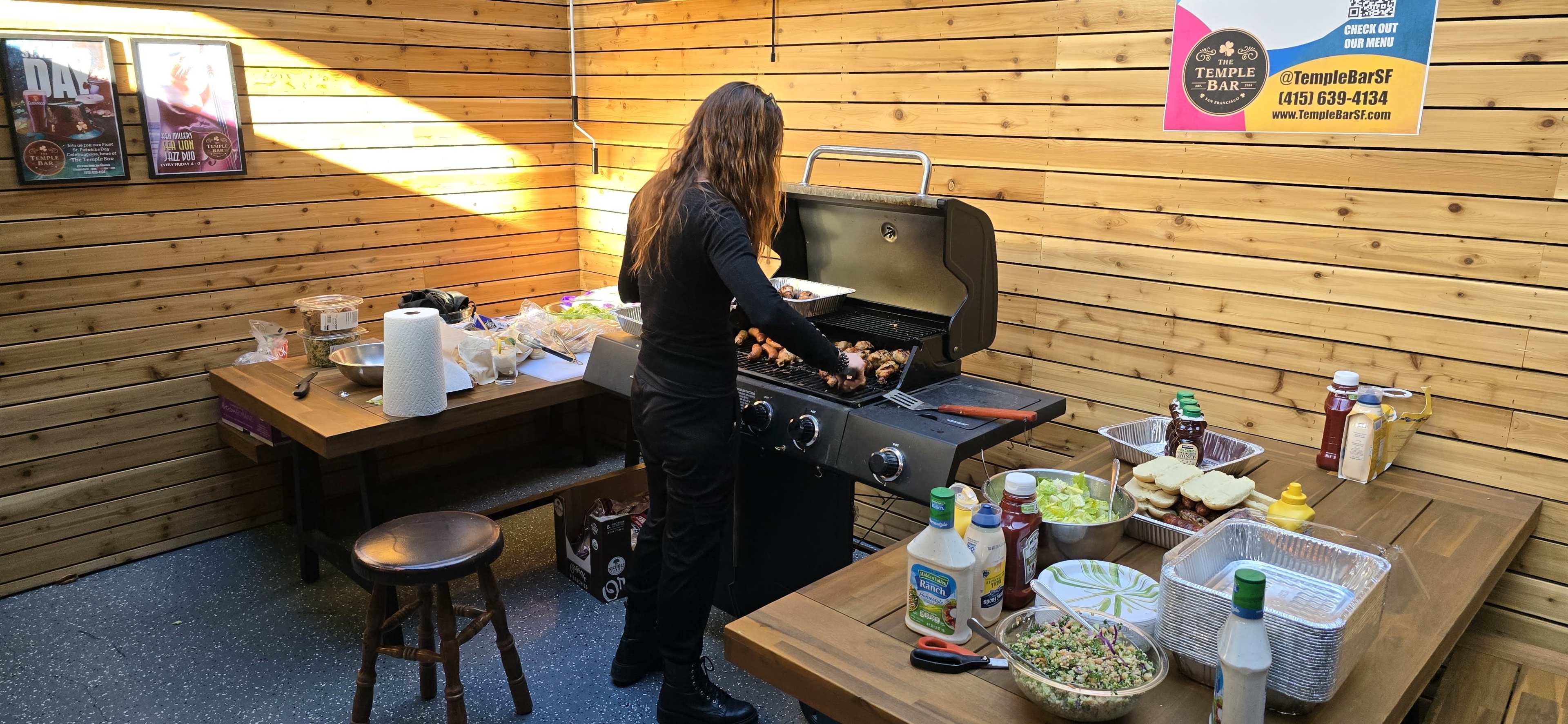 A person cooks on a grill in a wooden outdoor kitchen, surrounded by tables with various condiments and food items.
