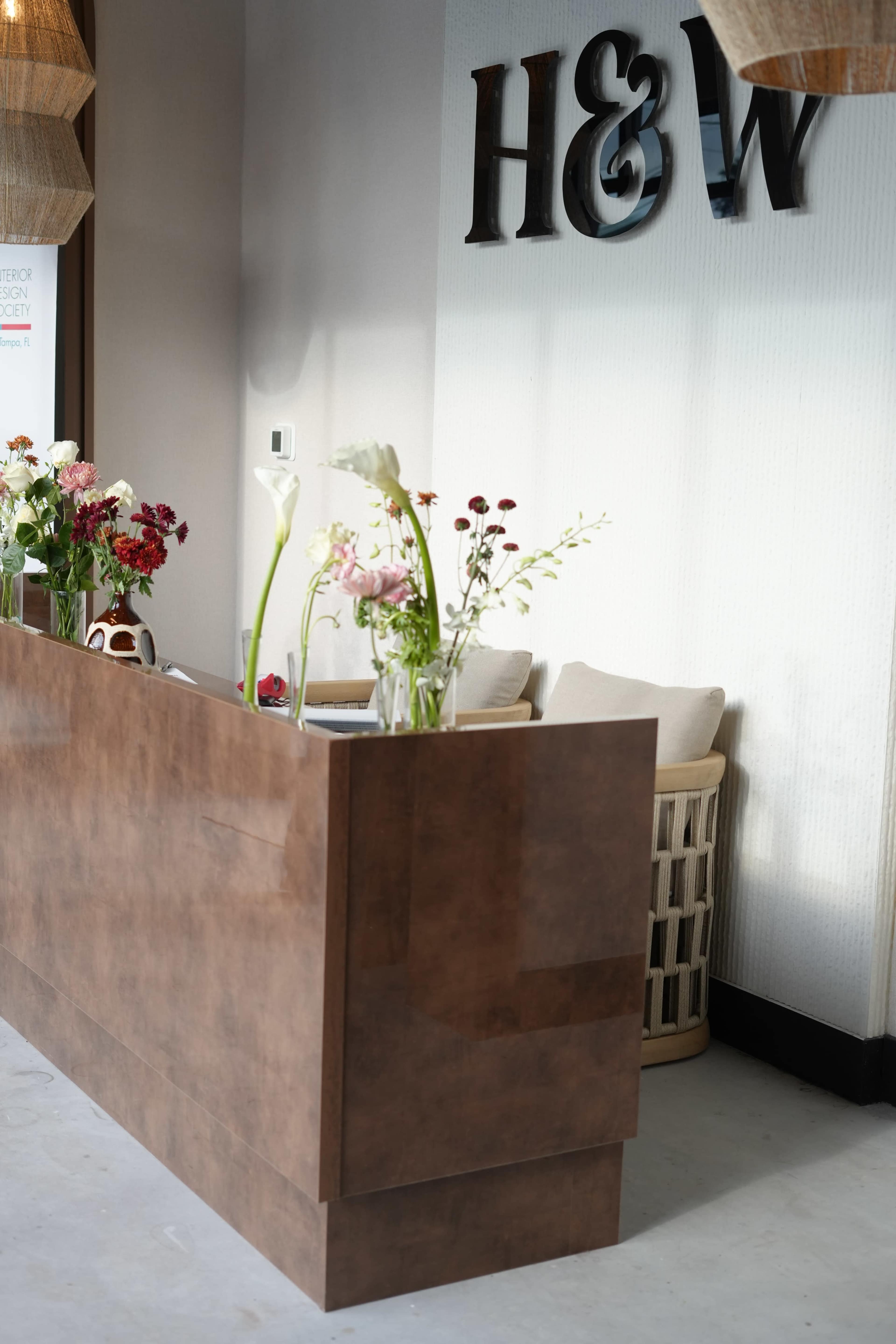The image shows a modern reception desk with a variety of flowers arranged in vases on top, situated in a well-lit space.