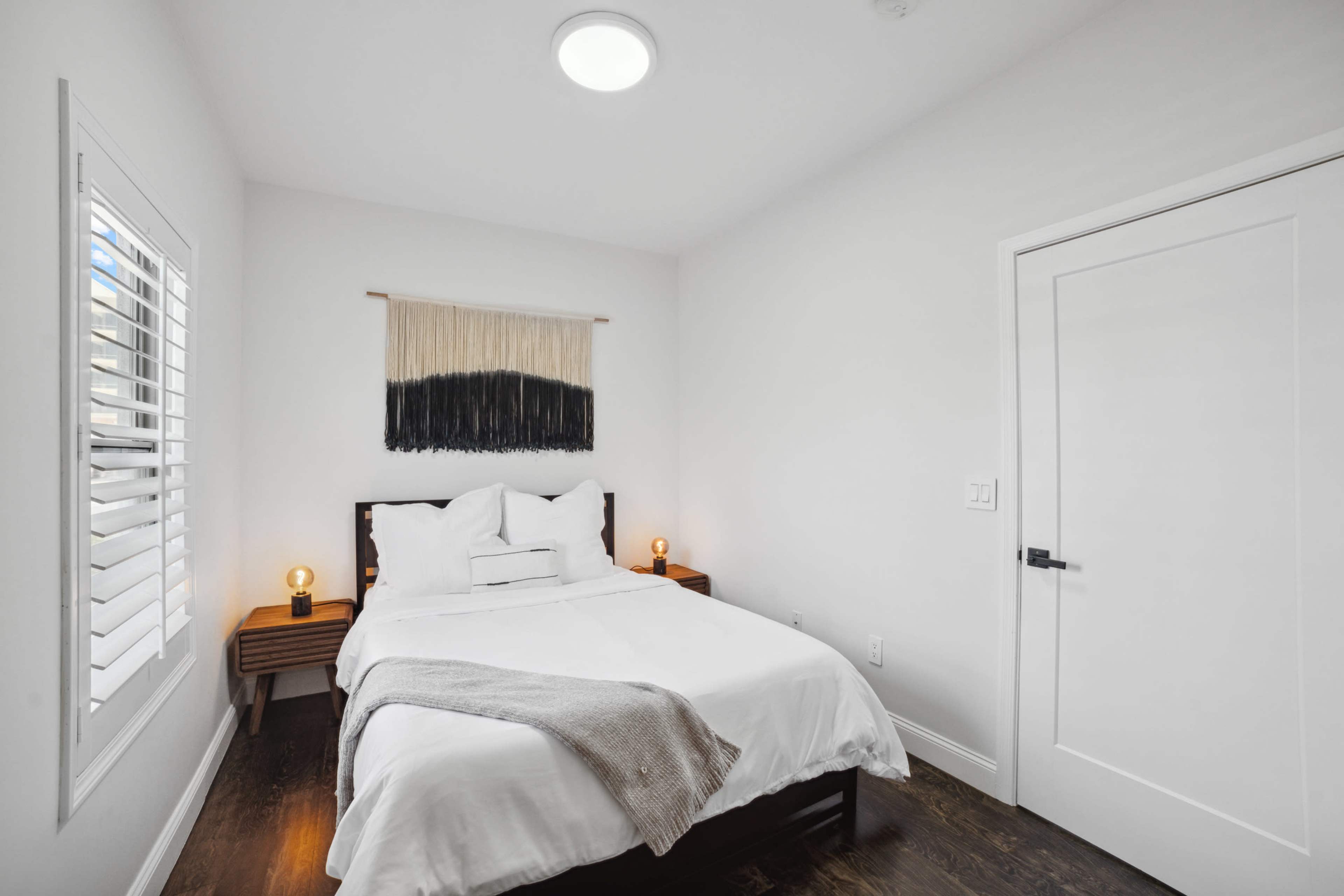 A bright bedroom with a white bedspread, bedside tables with lamps, and a wall hanging above the bed.