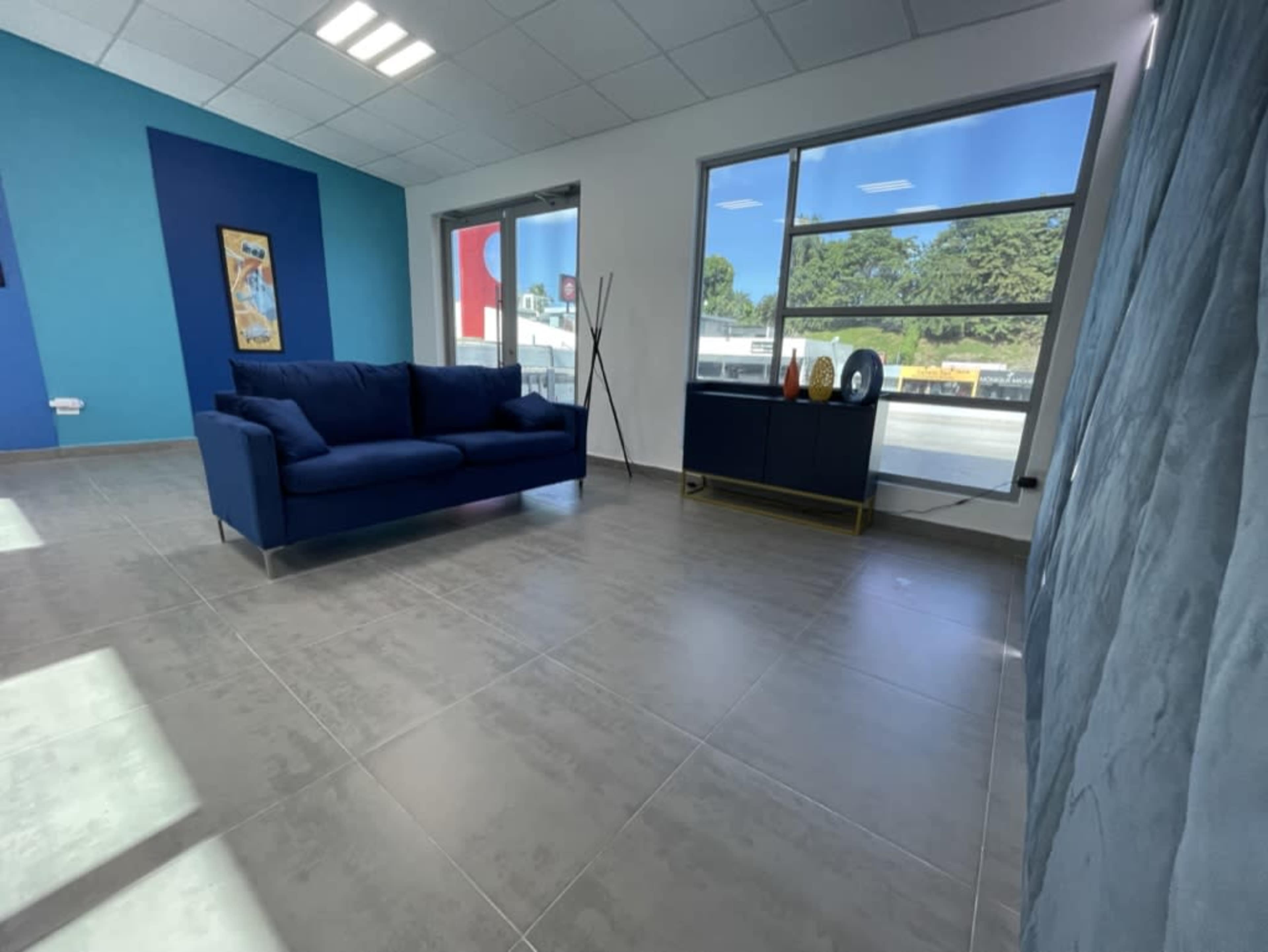 The image shows a modern living room with a blue couch, a console table, and large windows letting in daylight.