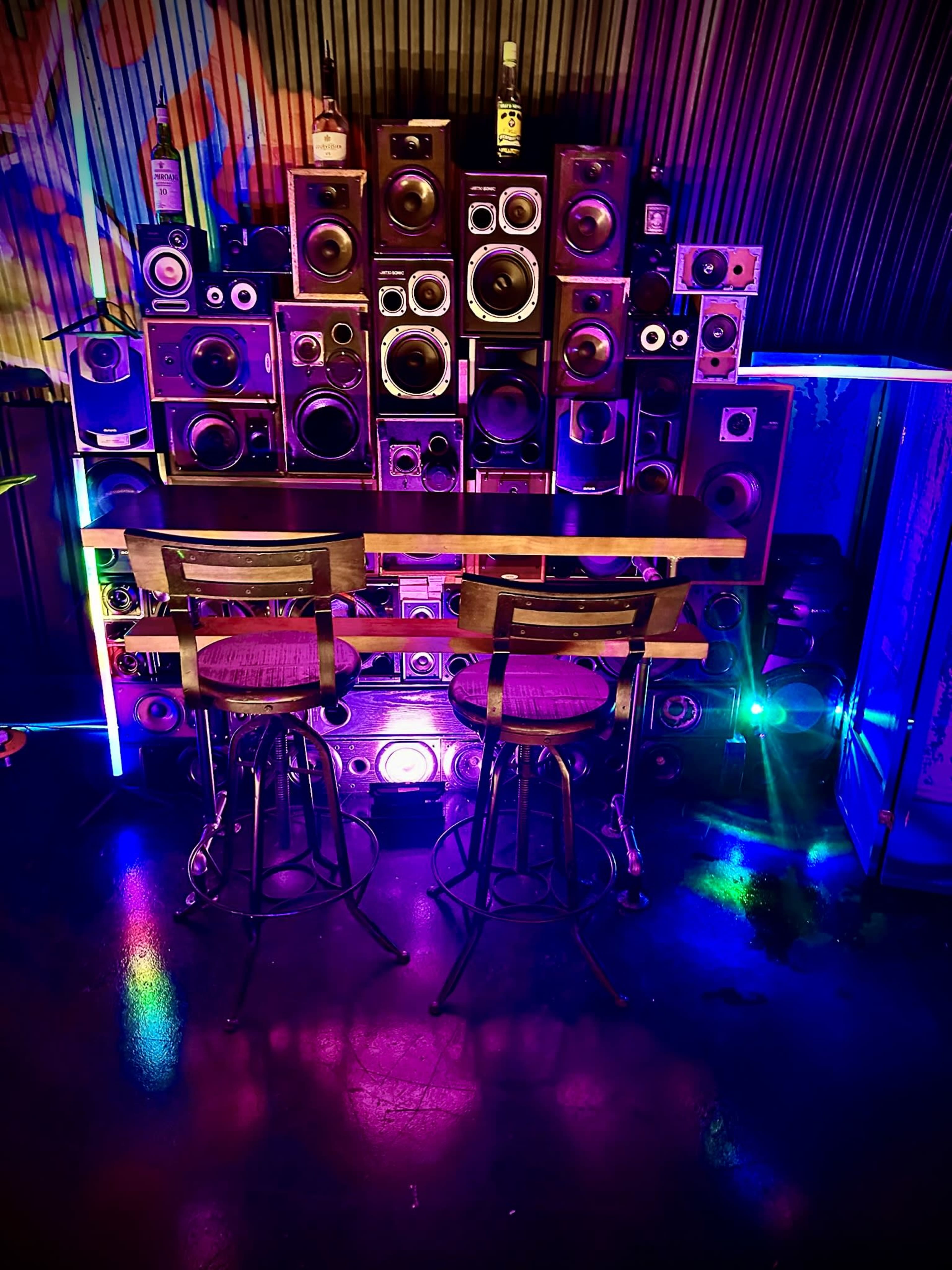The image shows a bar area with two stools in front of a wall of speakers, illuminated by colorful lights.