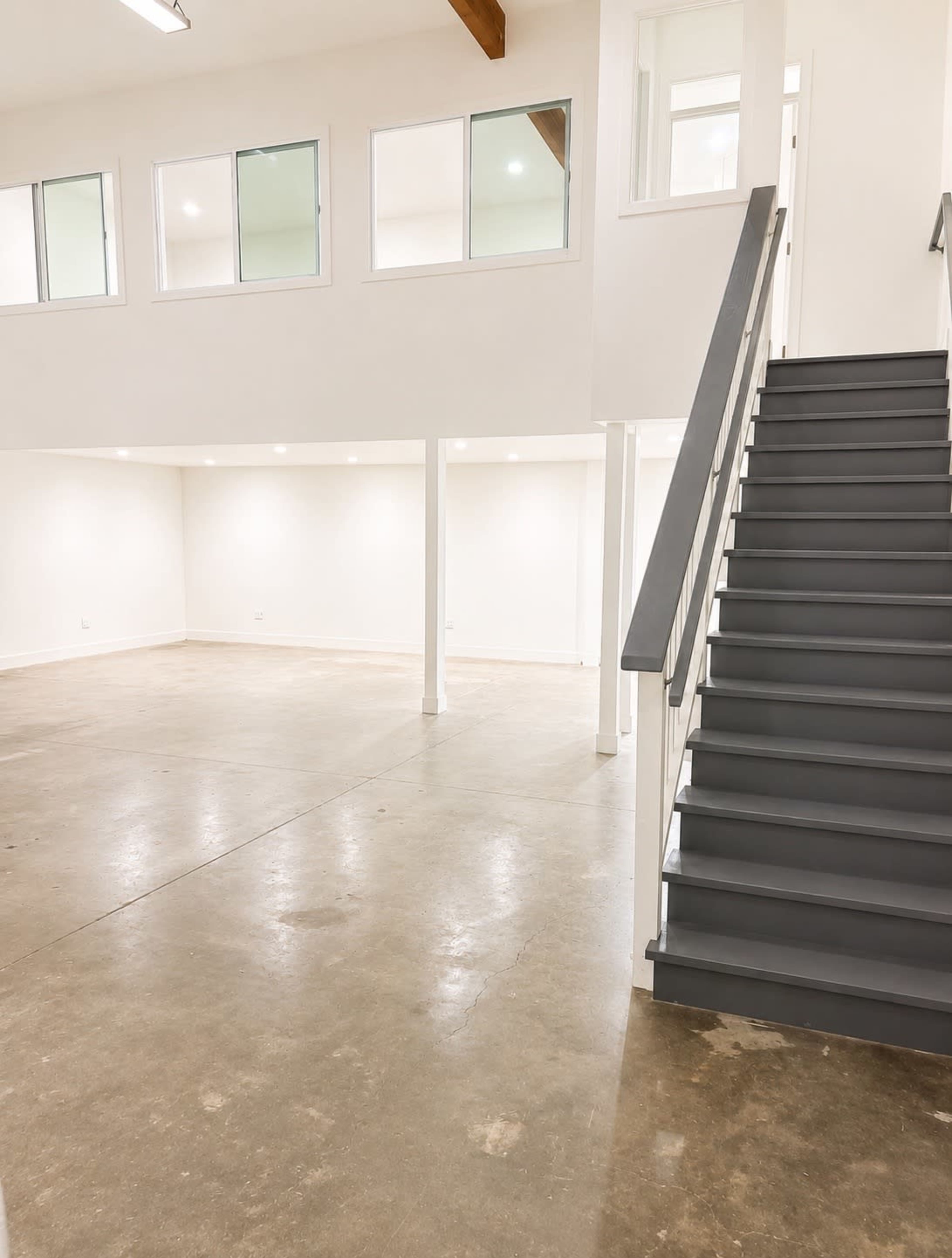 The image shows an empty, well-lit interior space featuring concrete flooring, white walls, and a staircase leading to an upper level.