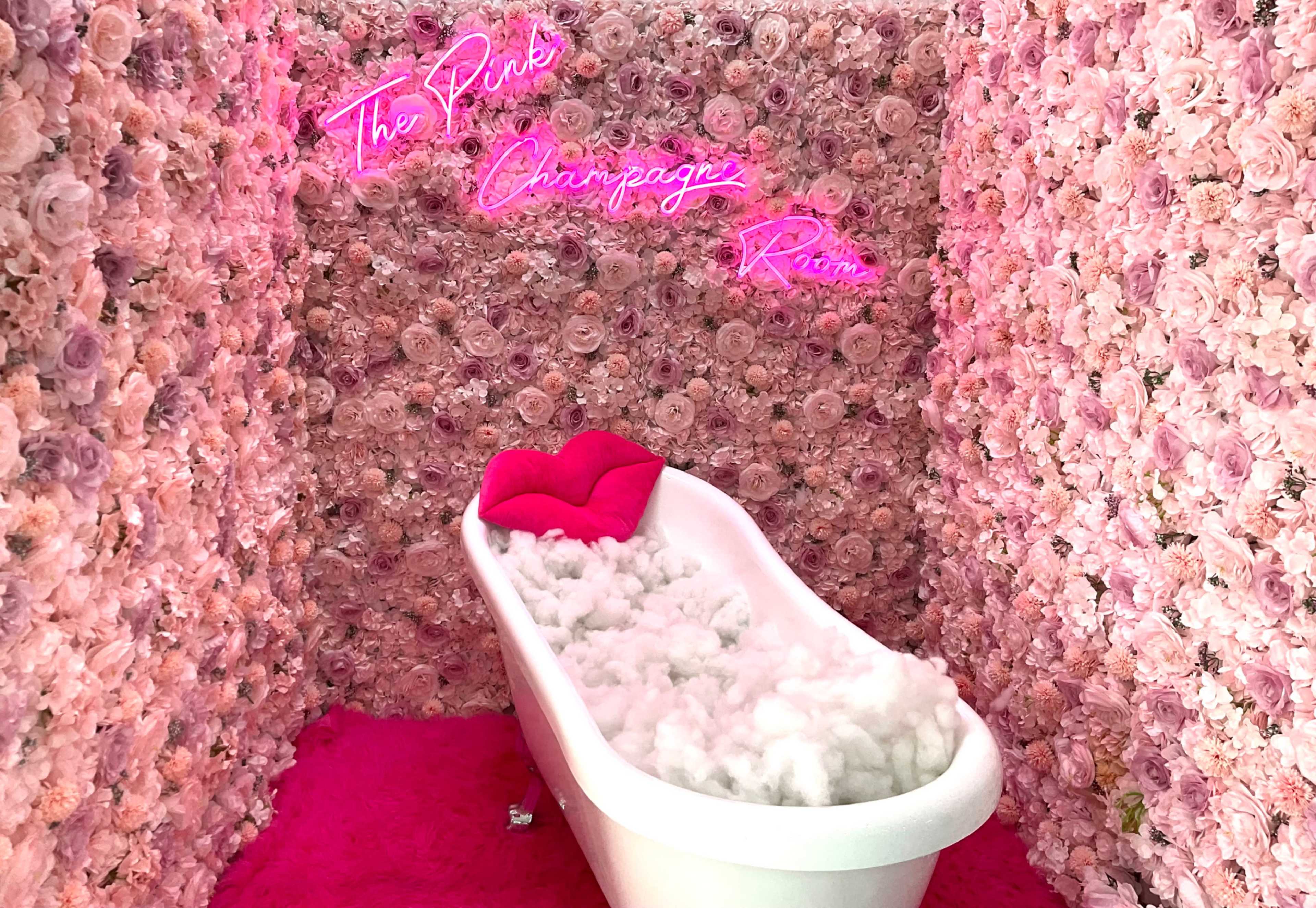 The scene features a white bathtub filled with cotton fluff, surrounded by a wall of pink flowers and illuminated by neon signs that read "The Pink Champagne Room."
