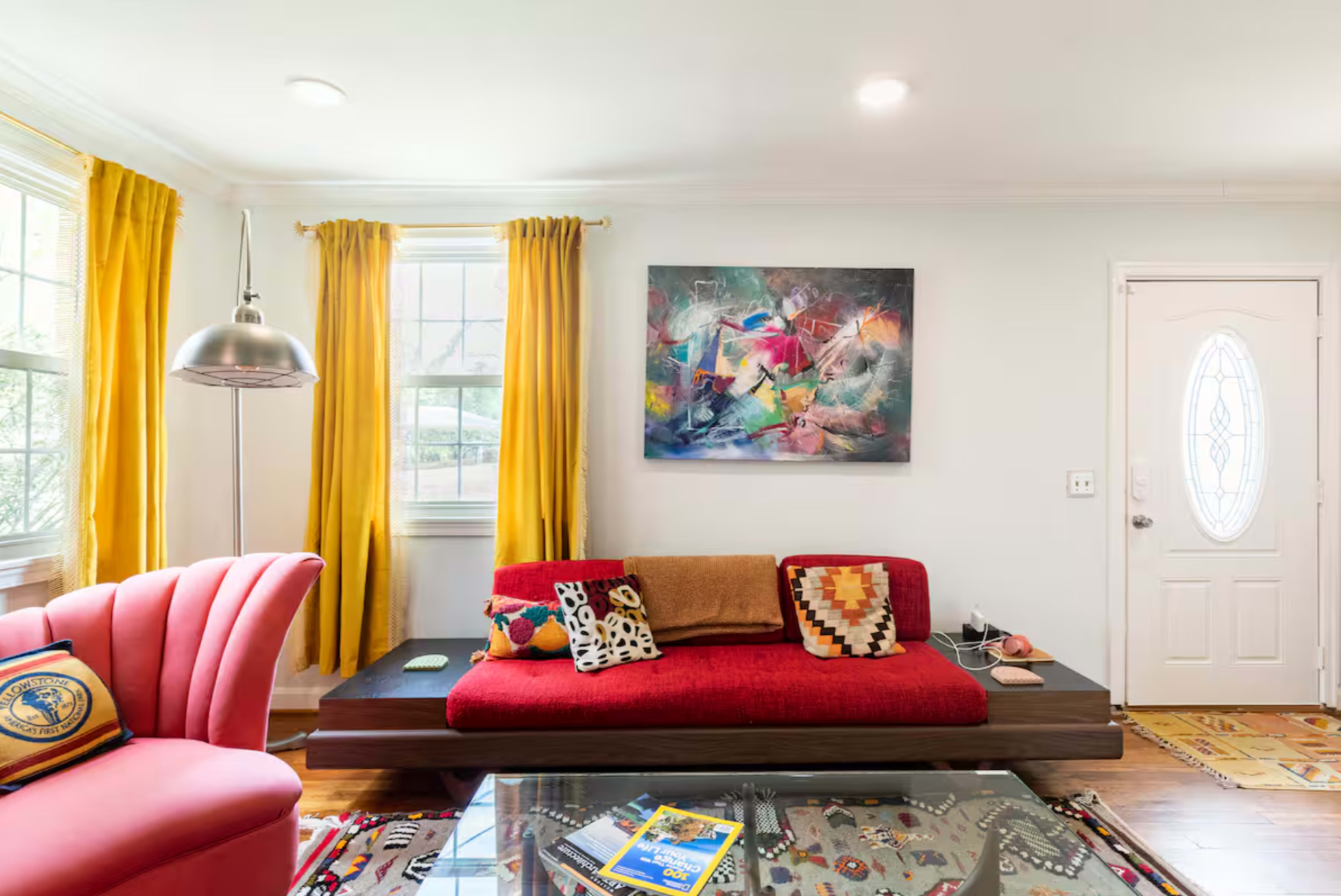 The image shows a living room with a red sofa, a pink chair, a glass coffee table, vibrant yellow curtains, and a colorful abstract painting on the wall.