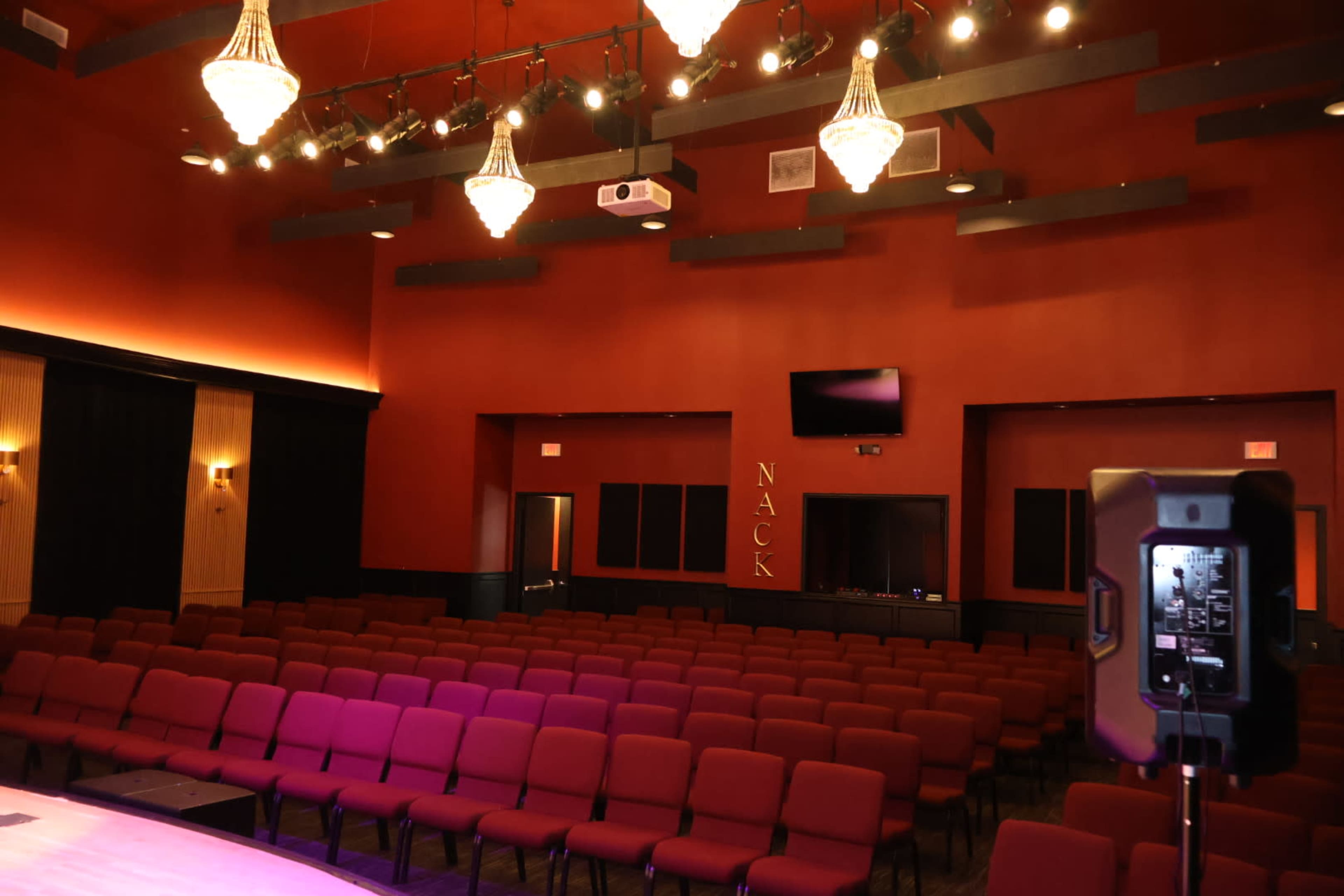 The image shows a theater interior featuring rows of red seats, a stage area with lighting fixtures, and a control station in the background.