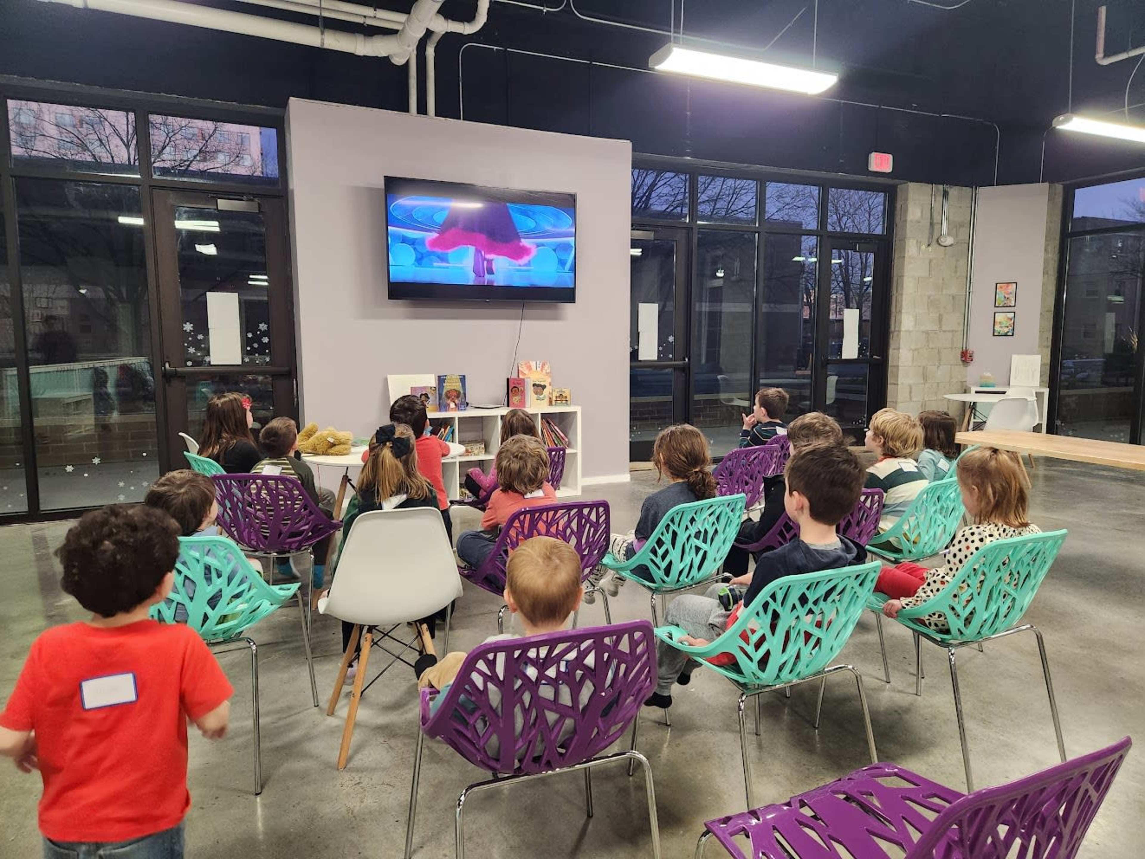 A group of children sits in colorful chairs, watching a video on a wall-mounted screen in a well-lit, modern room.