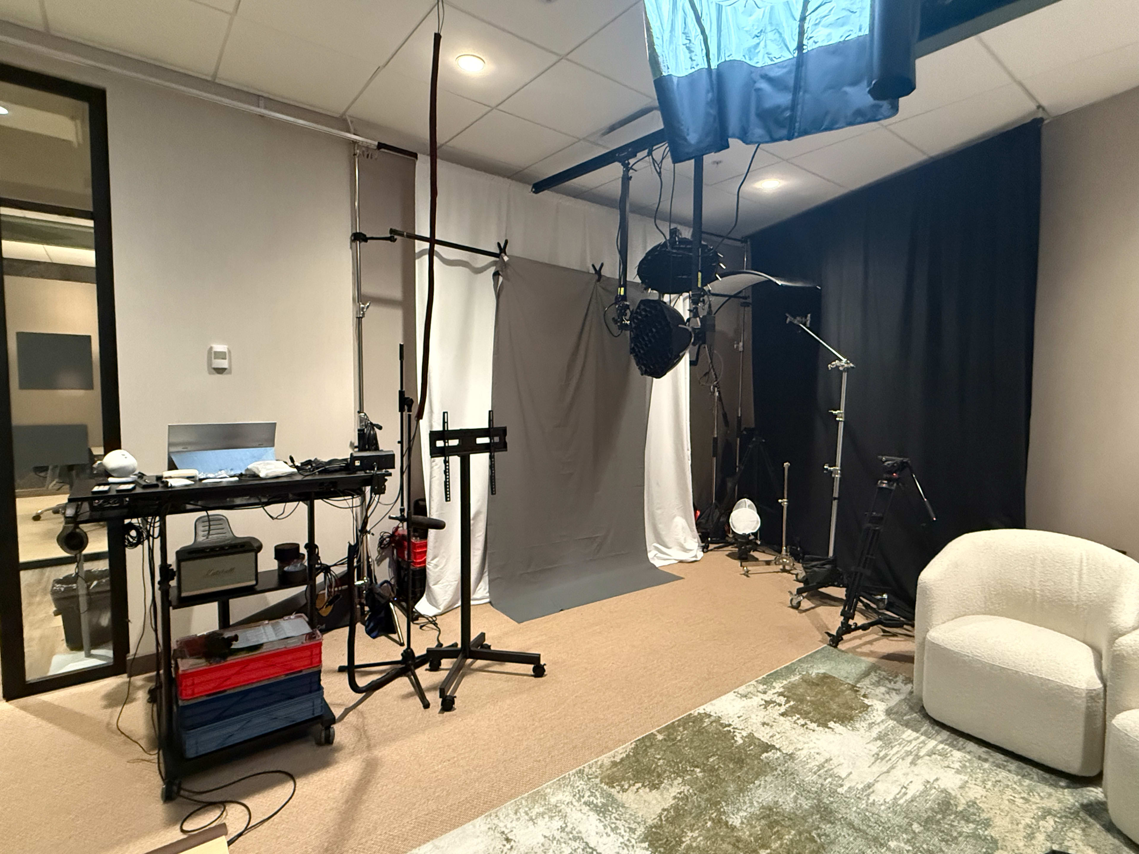 The image shows a photo studio setup with lighting equipment, a gray backdrop, and a few pieces of furniture.