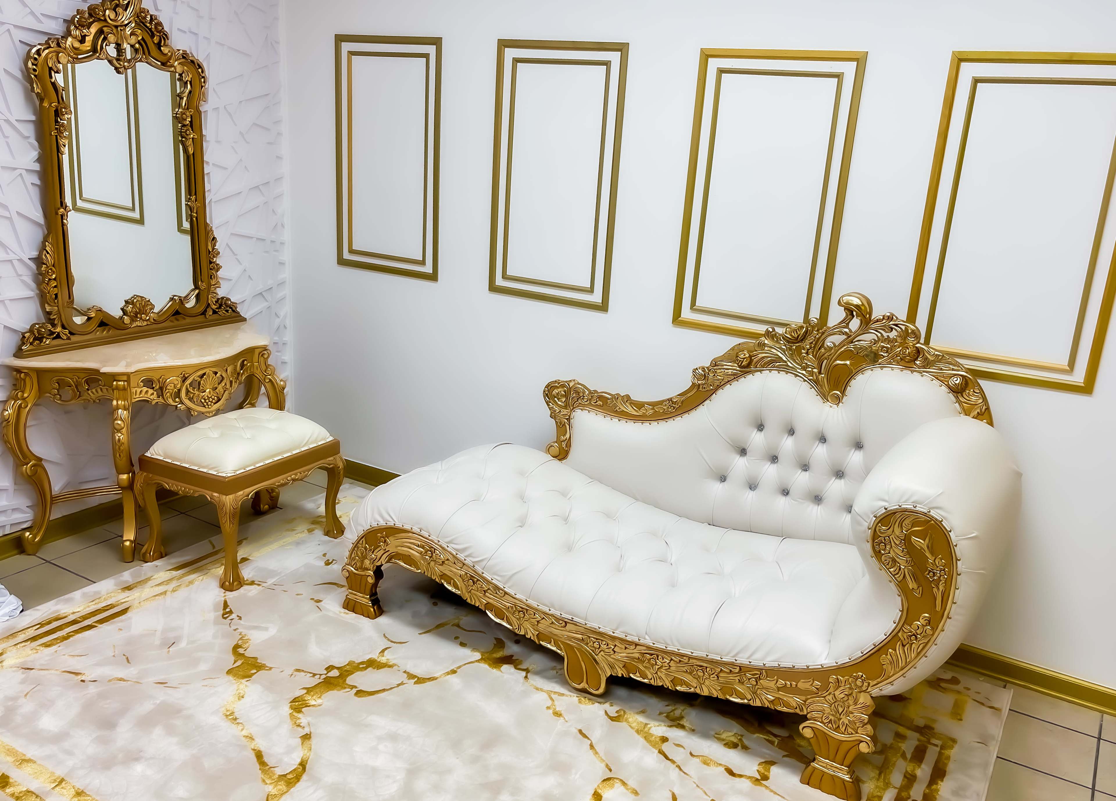A lavishly decorated room features a white tufted chaise lounge with gold accents, accompanied by a matching vanity and a gold-framed mirror, all set against a wall with empty picture frames.