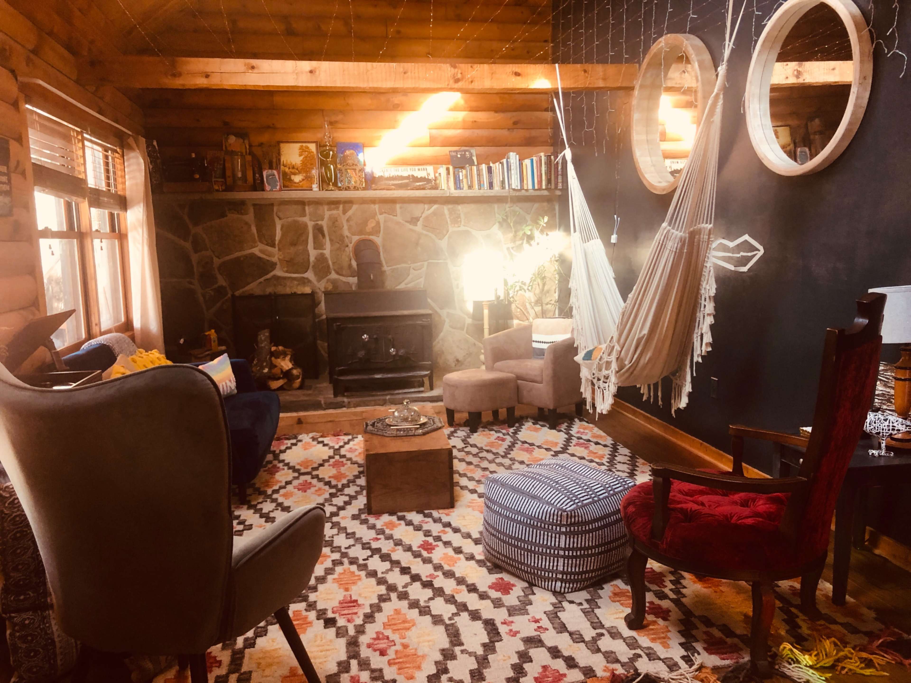 A cozy, rustic living room featuring a stone fireplace, a hammock, various seating options, and shelves lined with books.