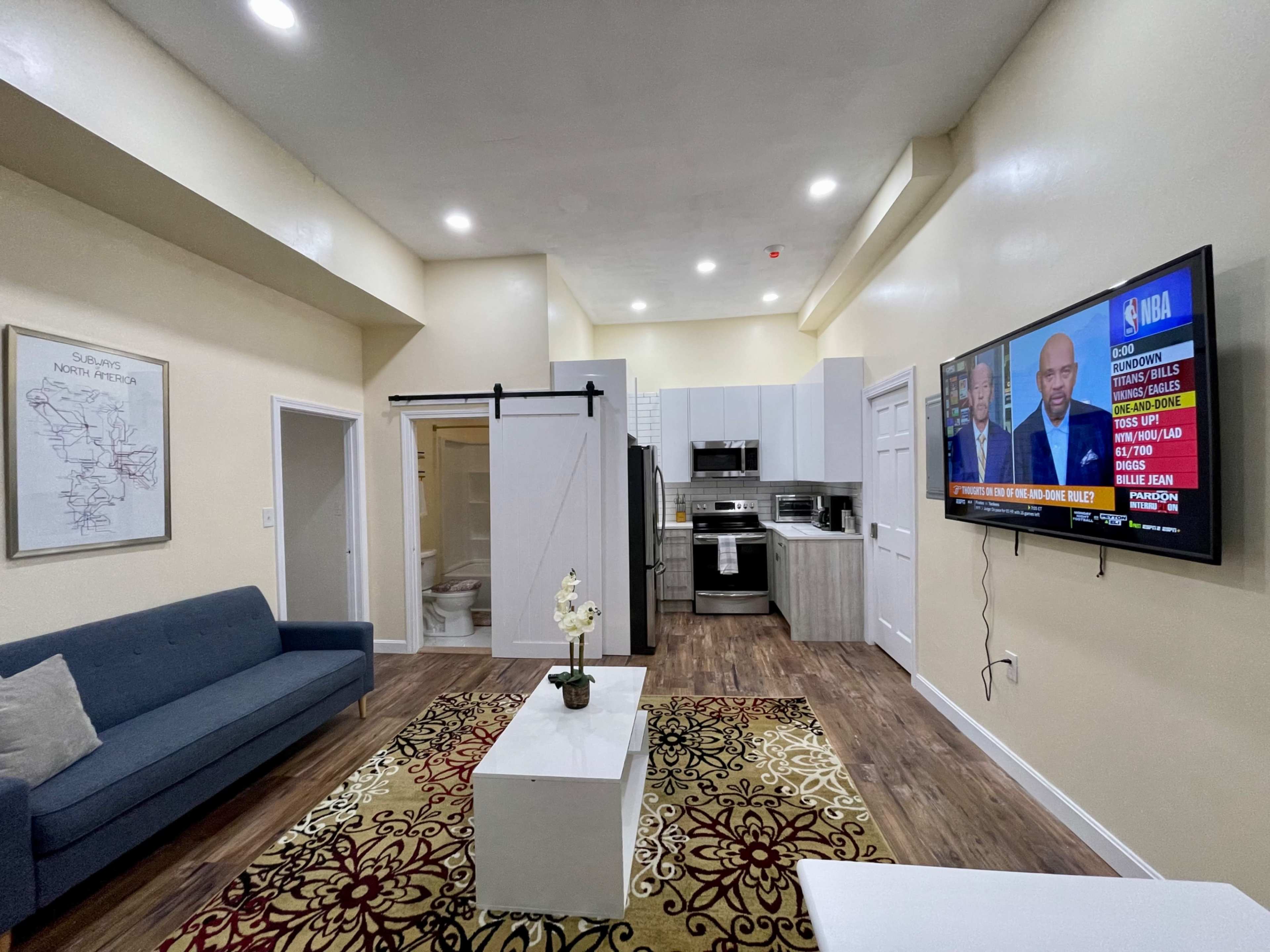 A modern living room with a sofa, a coffee table, a television mounted on the wall, and an adjacent kitchen area with a bathroom visible through a sliding door.