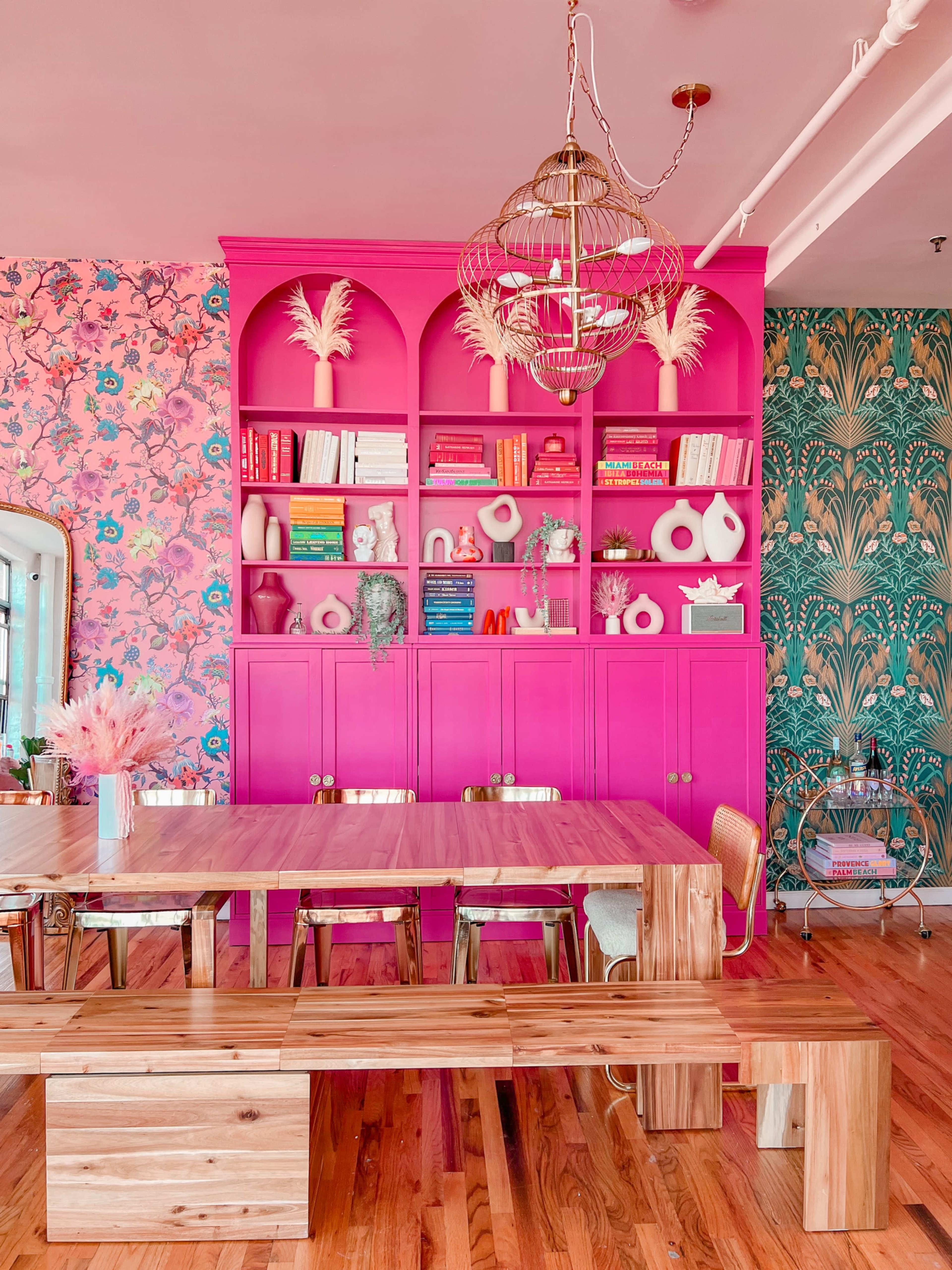 A brightly colored dining area features a pink bookshelf filled with decorative items and books, complemented by floral wallpaper and a wooden dining table with bench seating.