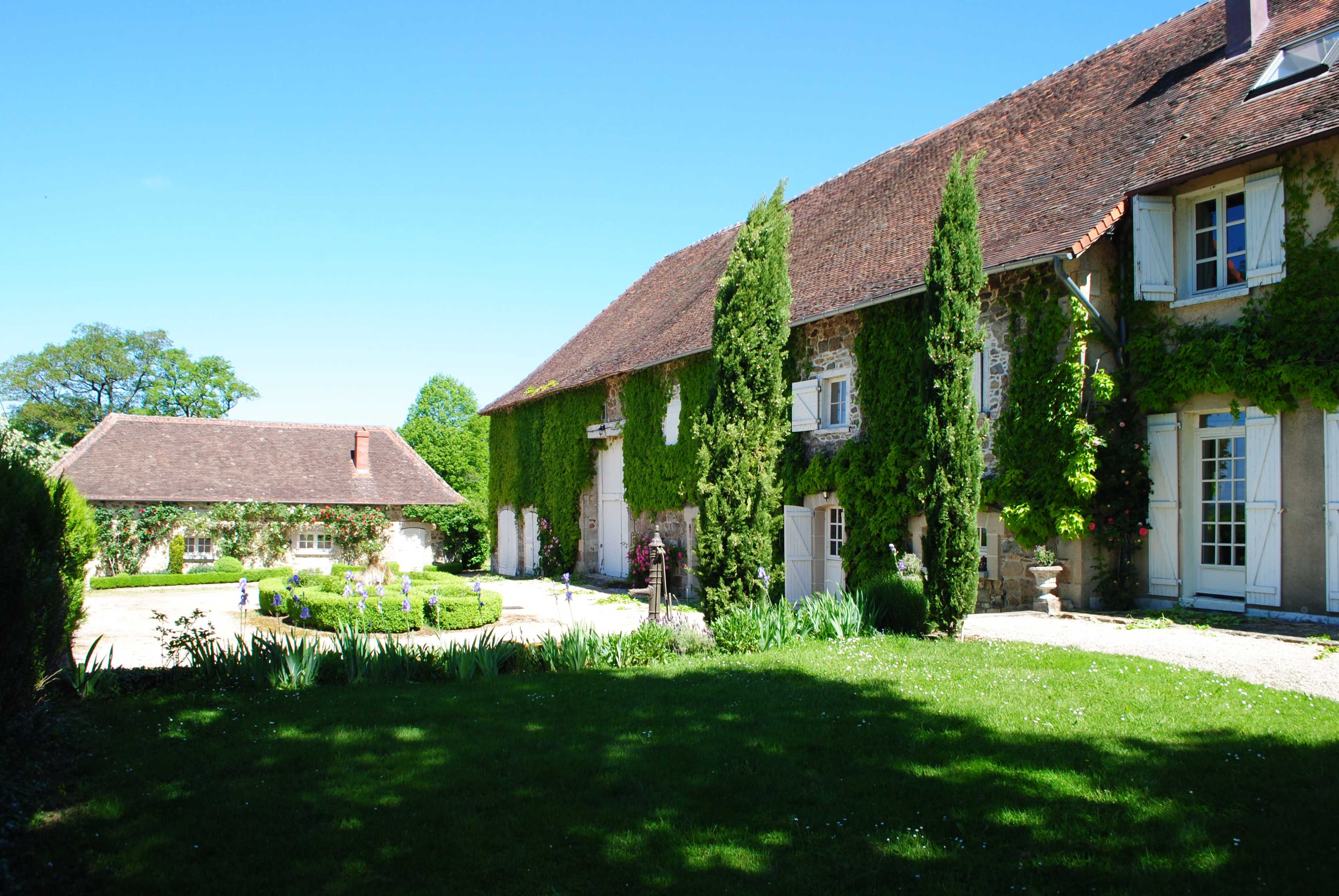 The image shows a charming stone farmhouse with a brown tiled roof, surrounded by lush greenery and a landscaped garden.