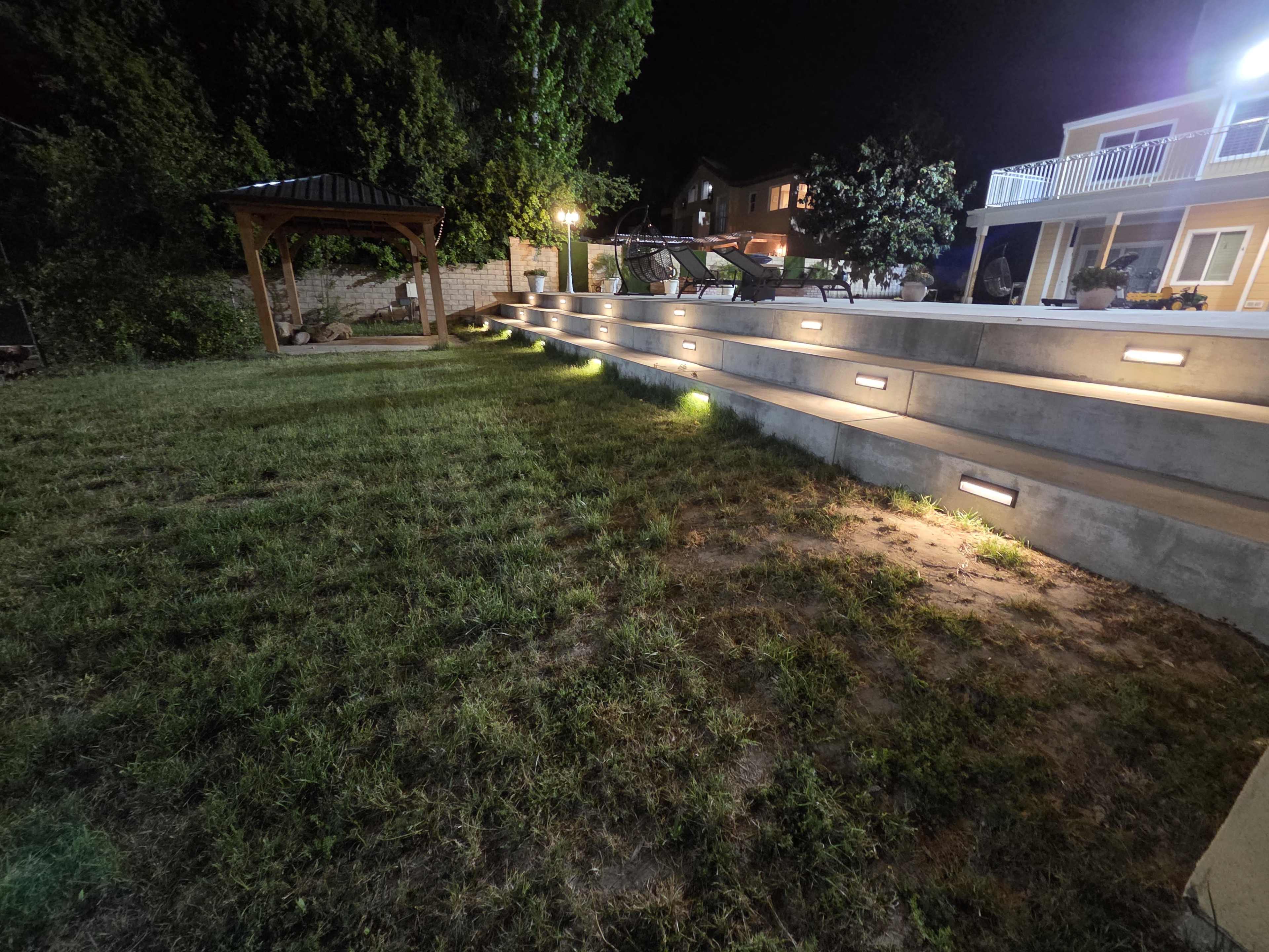 The image shows a backyard at night featuring illuminated steps that lead up to a landscaped area with a gazebo and a house in the background.