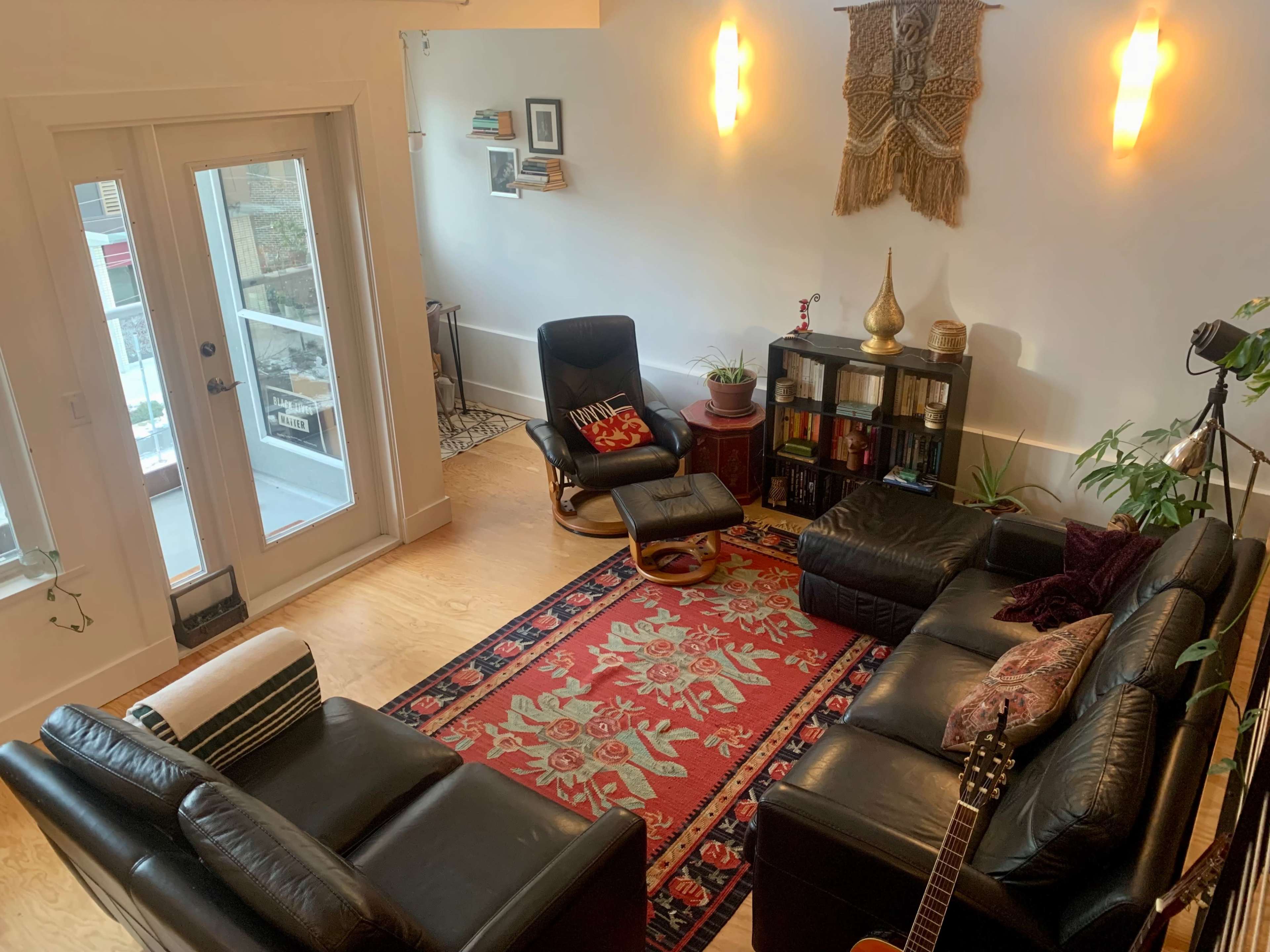 A cozy living room with black leather sofas, a patterned rug, and large windows that allow natural light.