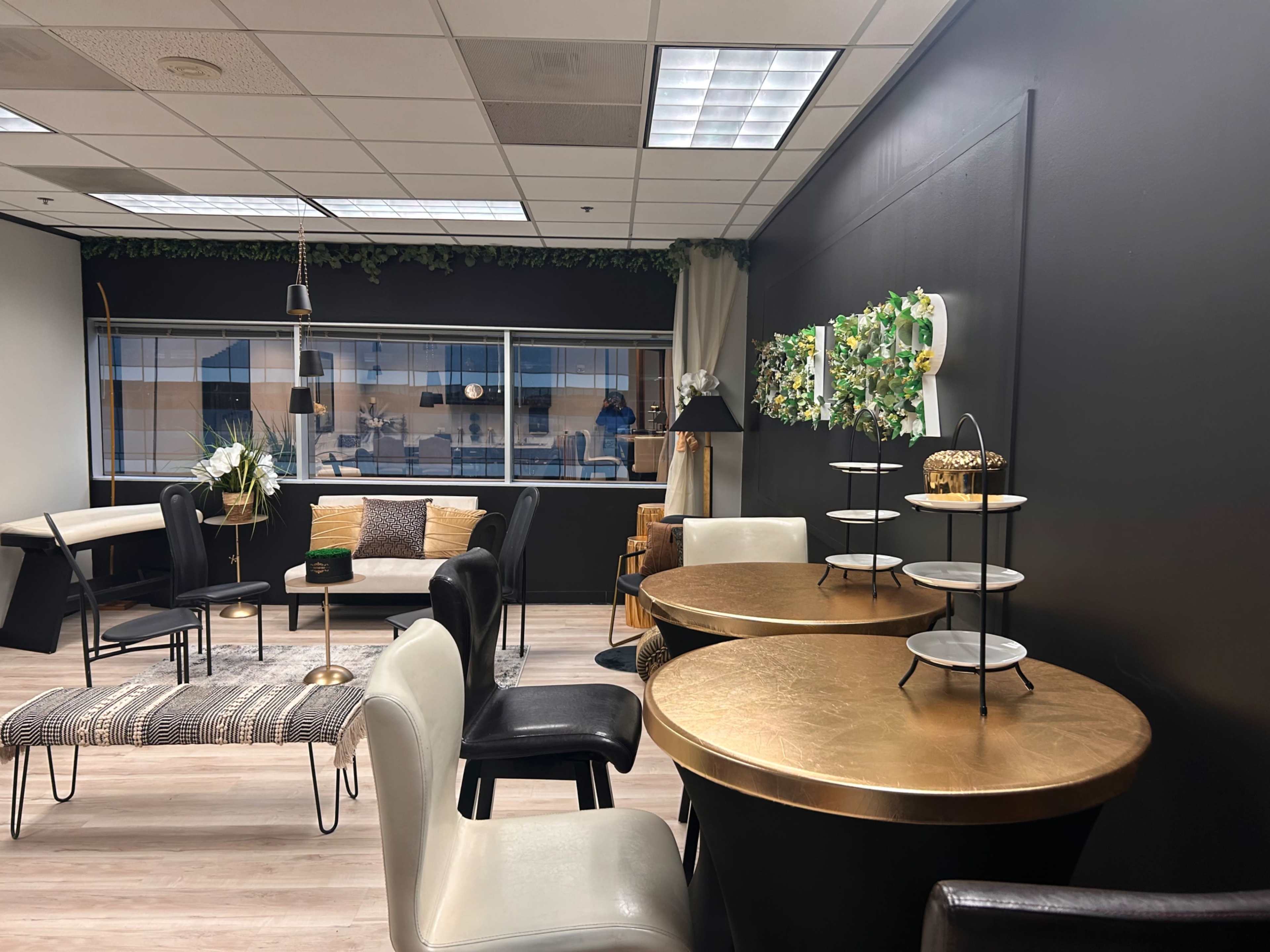 The image shows a stylish office space featuring a mix of seating arrangements, decorative plants, and round tables with a gold finish.