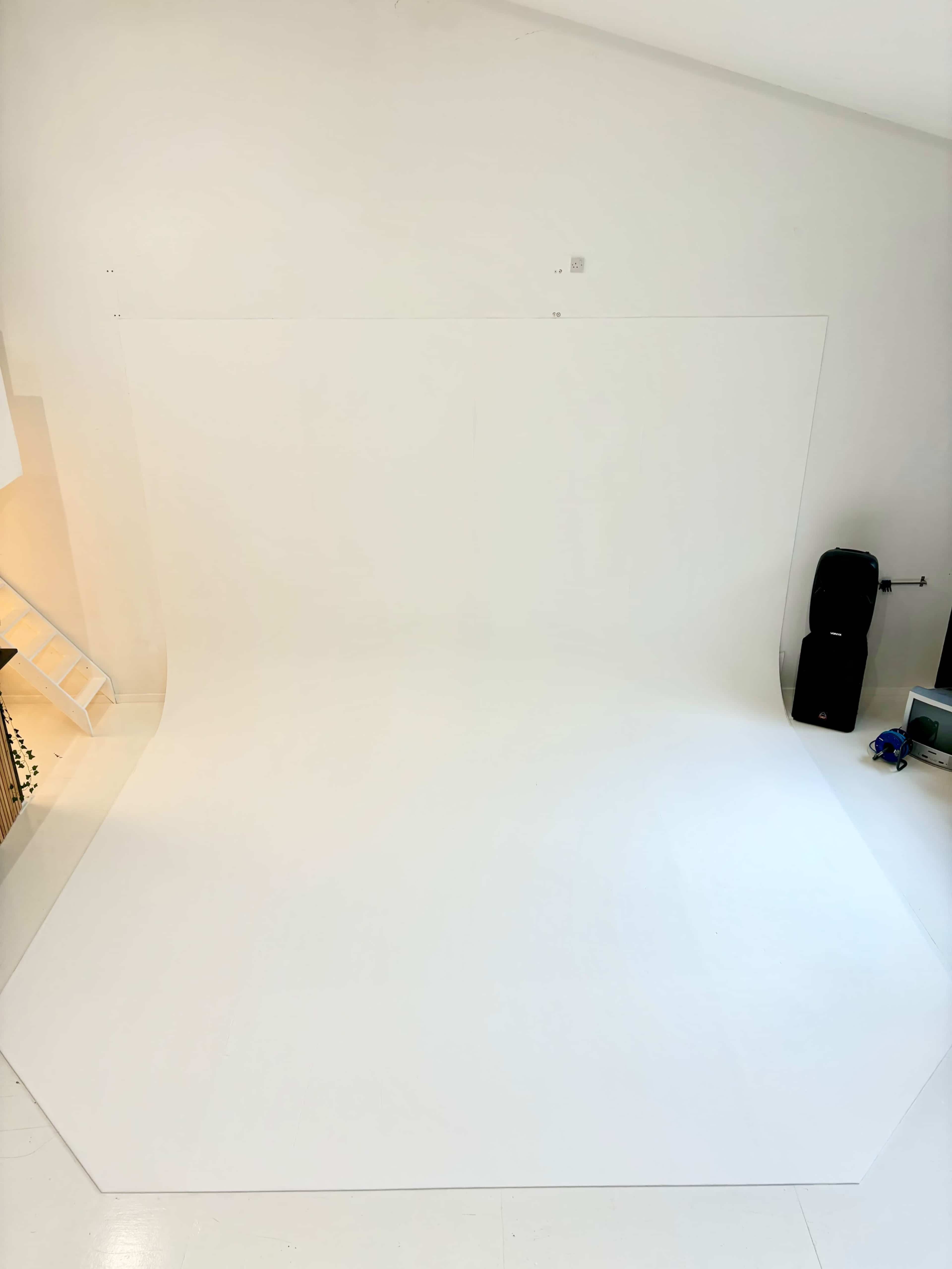 A spacious, blank white studio backdrop is set up in an empty room, with some reflective surfaces and equipment visible in the background.
