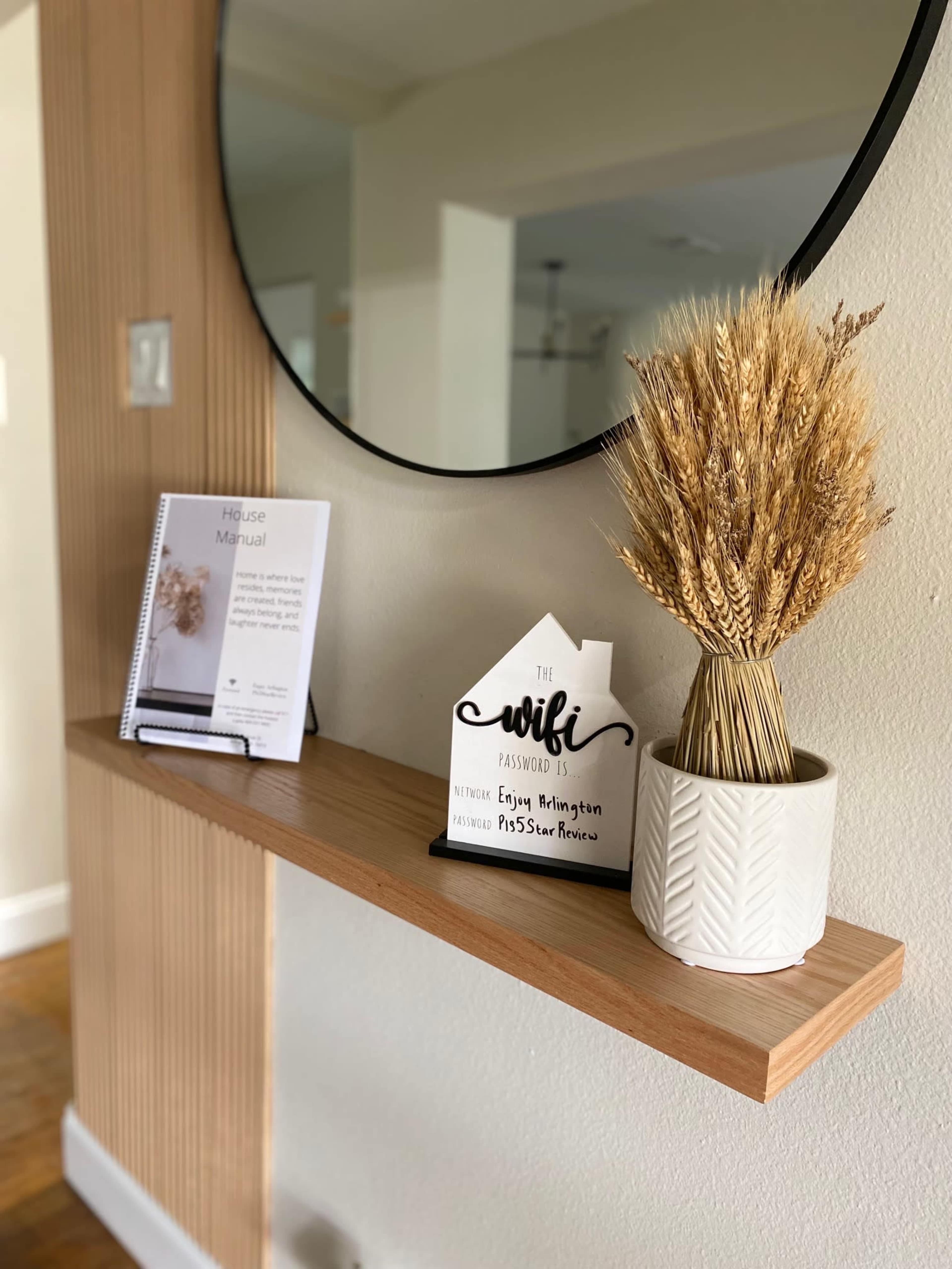 A circular mirror hangs above a wooden shelf that displays a vase of dried flowers and a house manual.