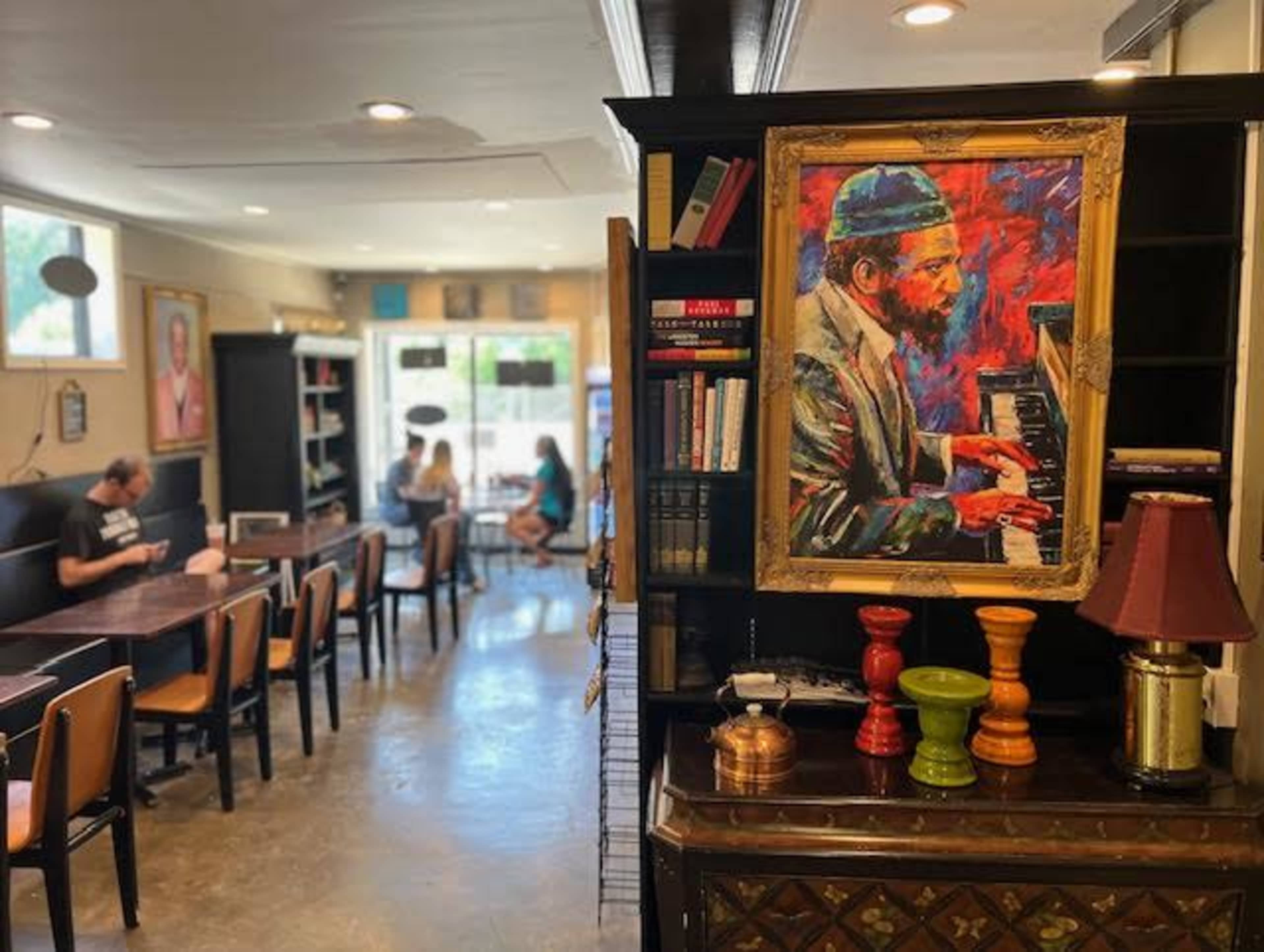The image shows an interior of a cafe featuring a colorful painting of a man playing piano, with shelves of books and patrons seated at tables in the background.