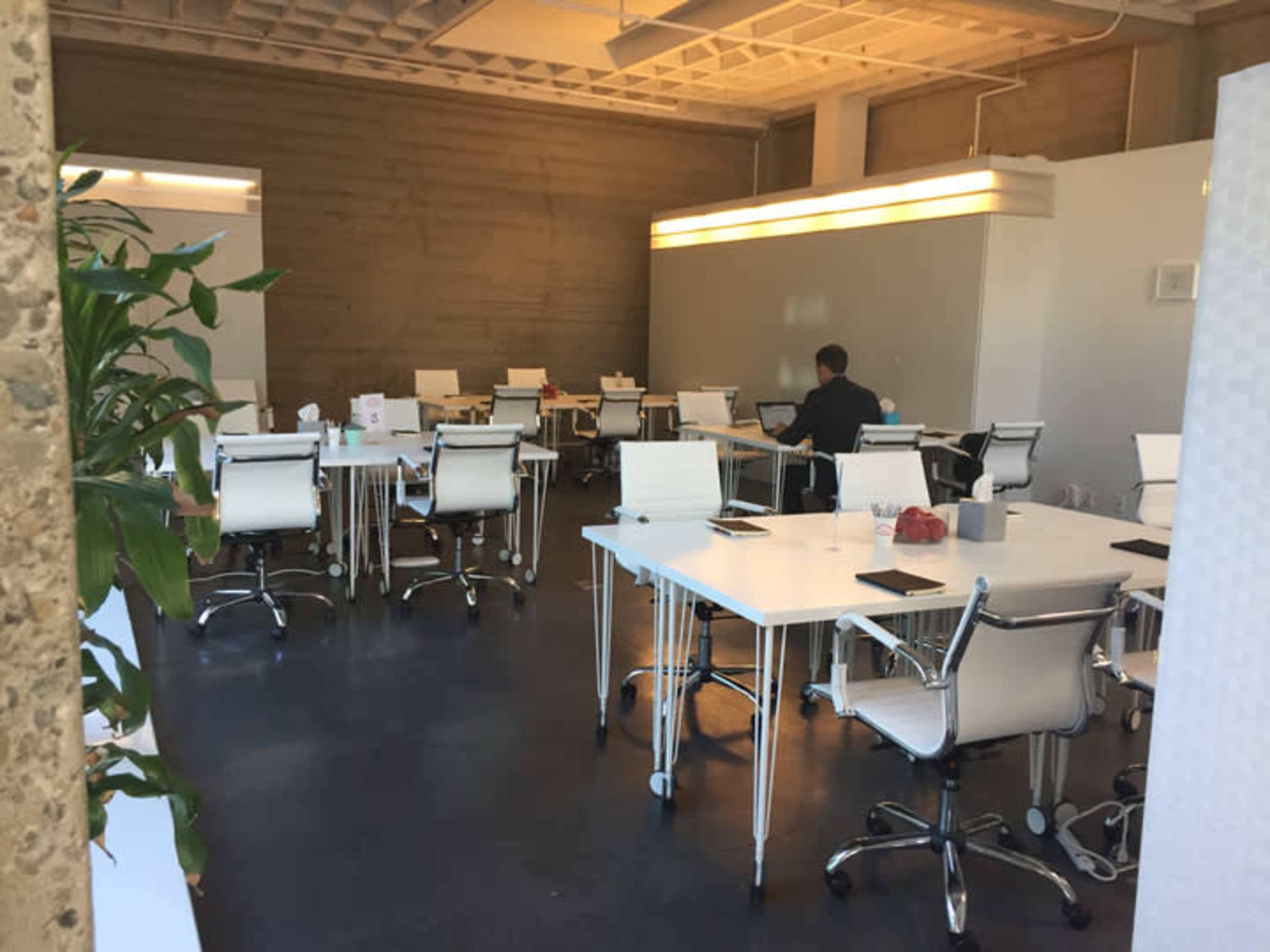 A modern office space features several white desks and chairs, with a lone person working at one of the tables in the background.
