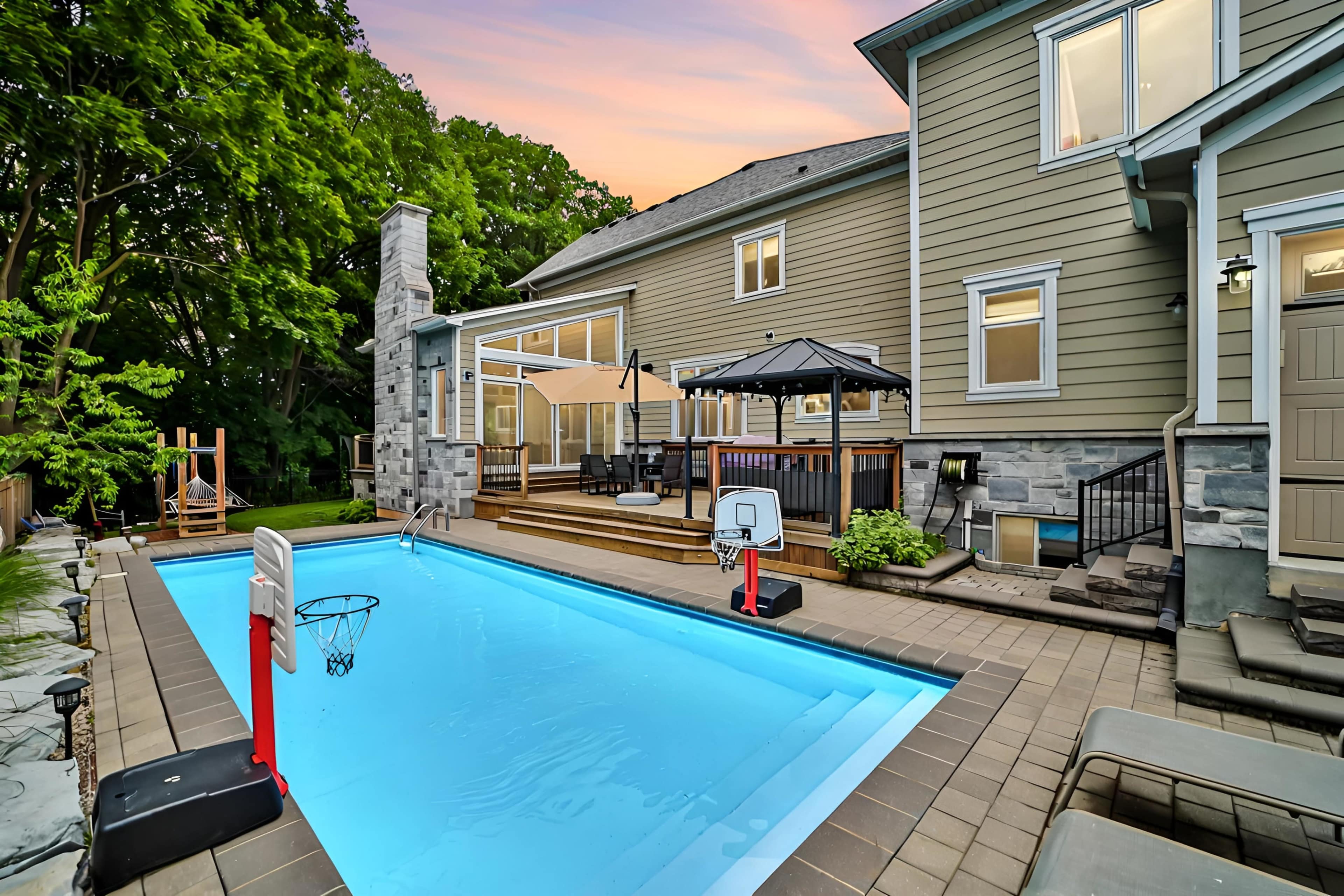 A backyard features a swimming pool with a basketball hoop, surrounded by lounge chairs and a patio with an umbrella and seating area.