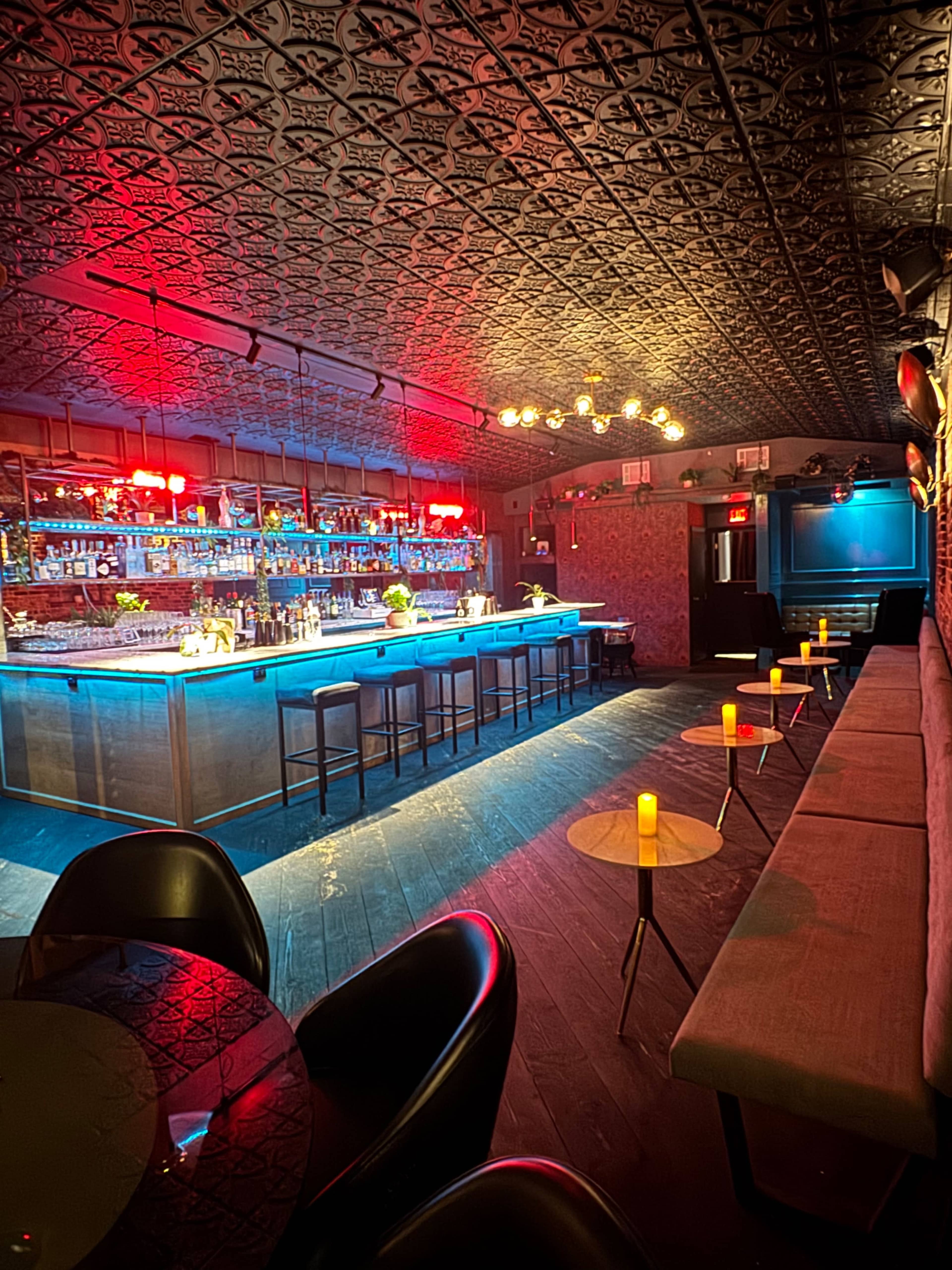 The image shows a stylish bar interior with a long illuminated counter, seating along the walls, and ambient lighting from candles on small tables.