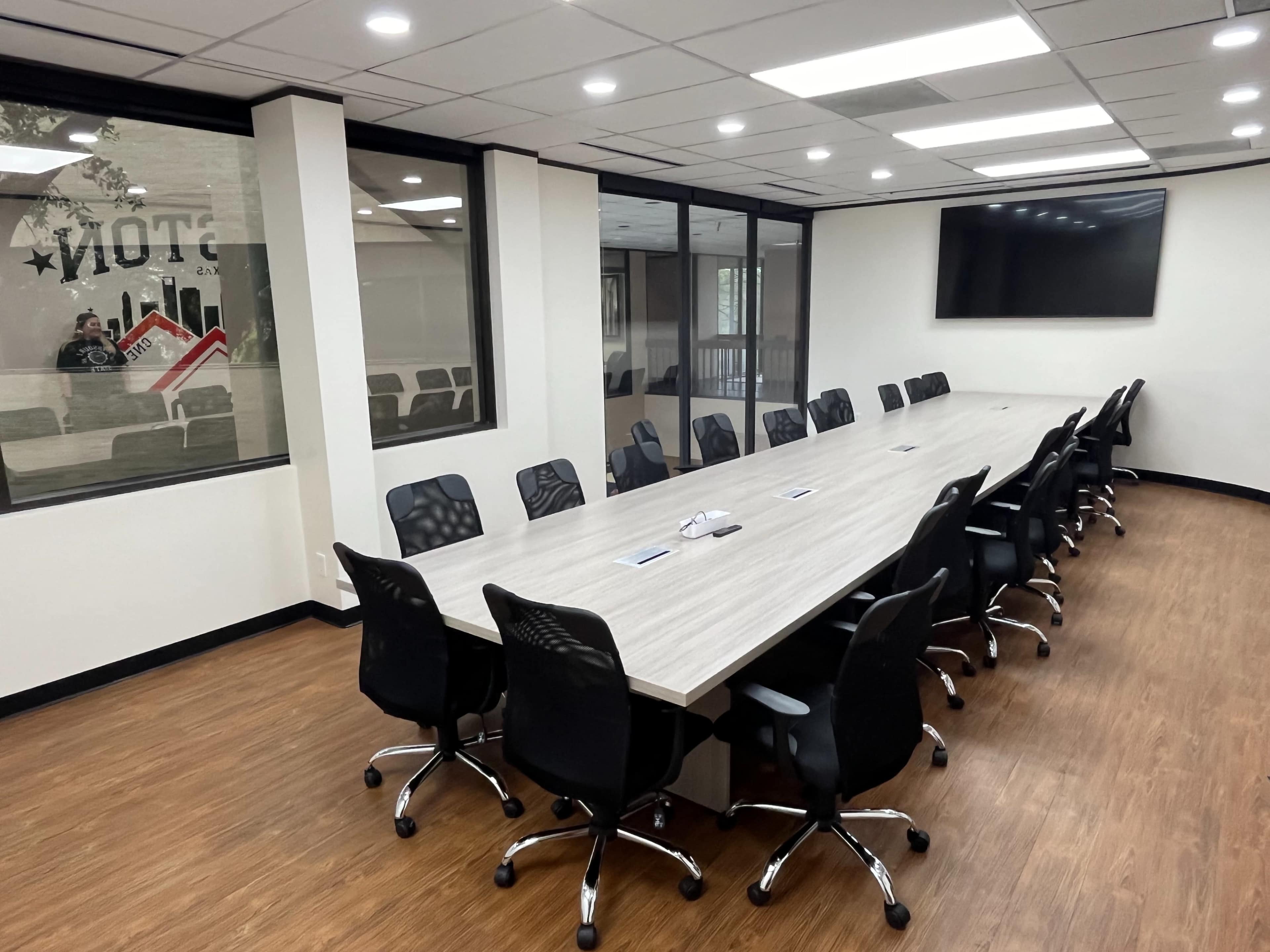A large conference room features a long table surrounded by black rolling chairs, with windows and a wall-mounted TV.