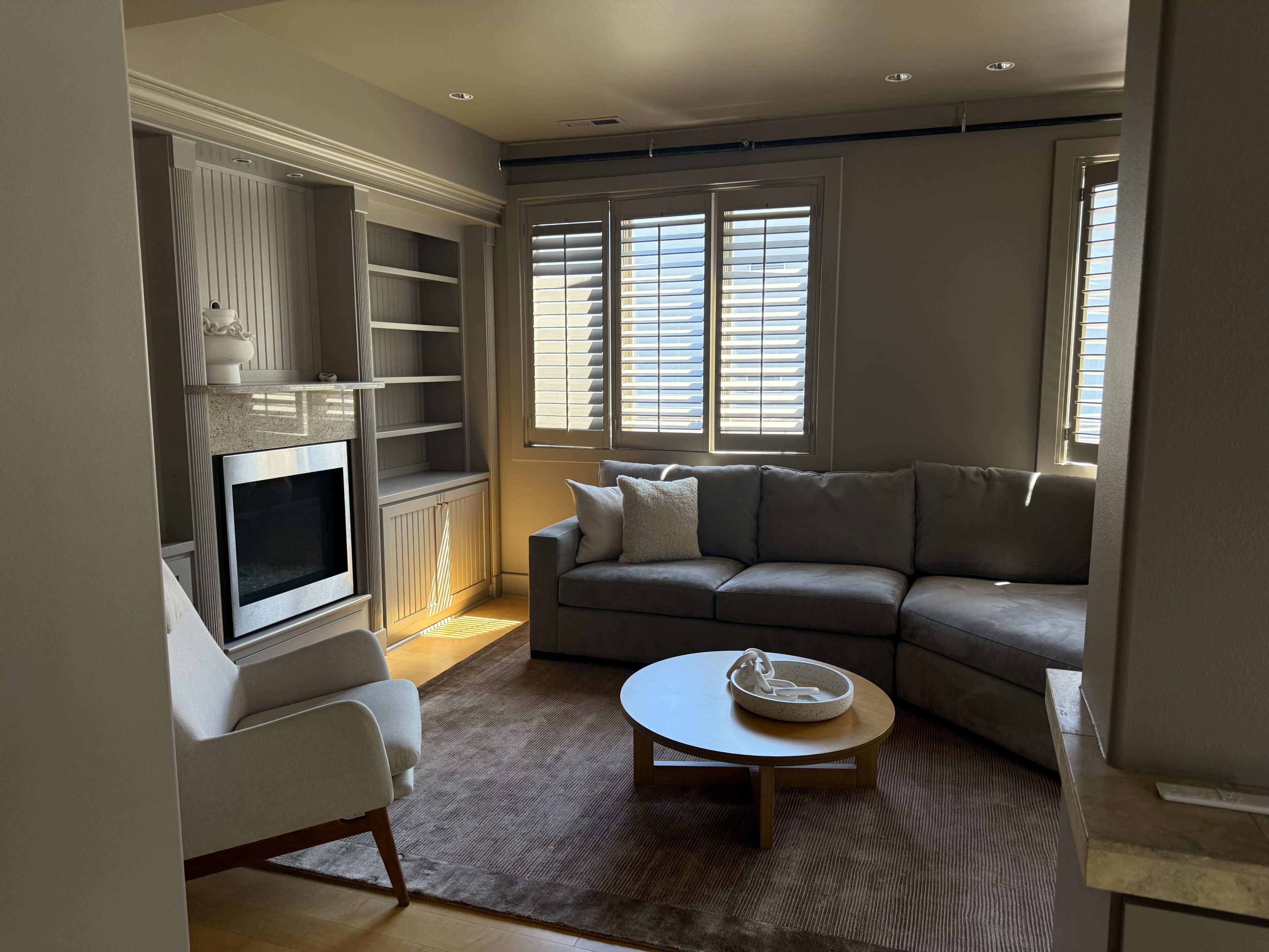 The image shows a living room with a gray sectional sofa, a round coffee table, a white armchair, and large windows with shutters letting in natural light.