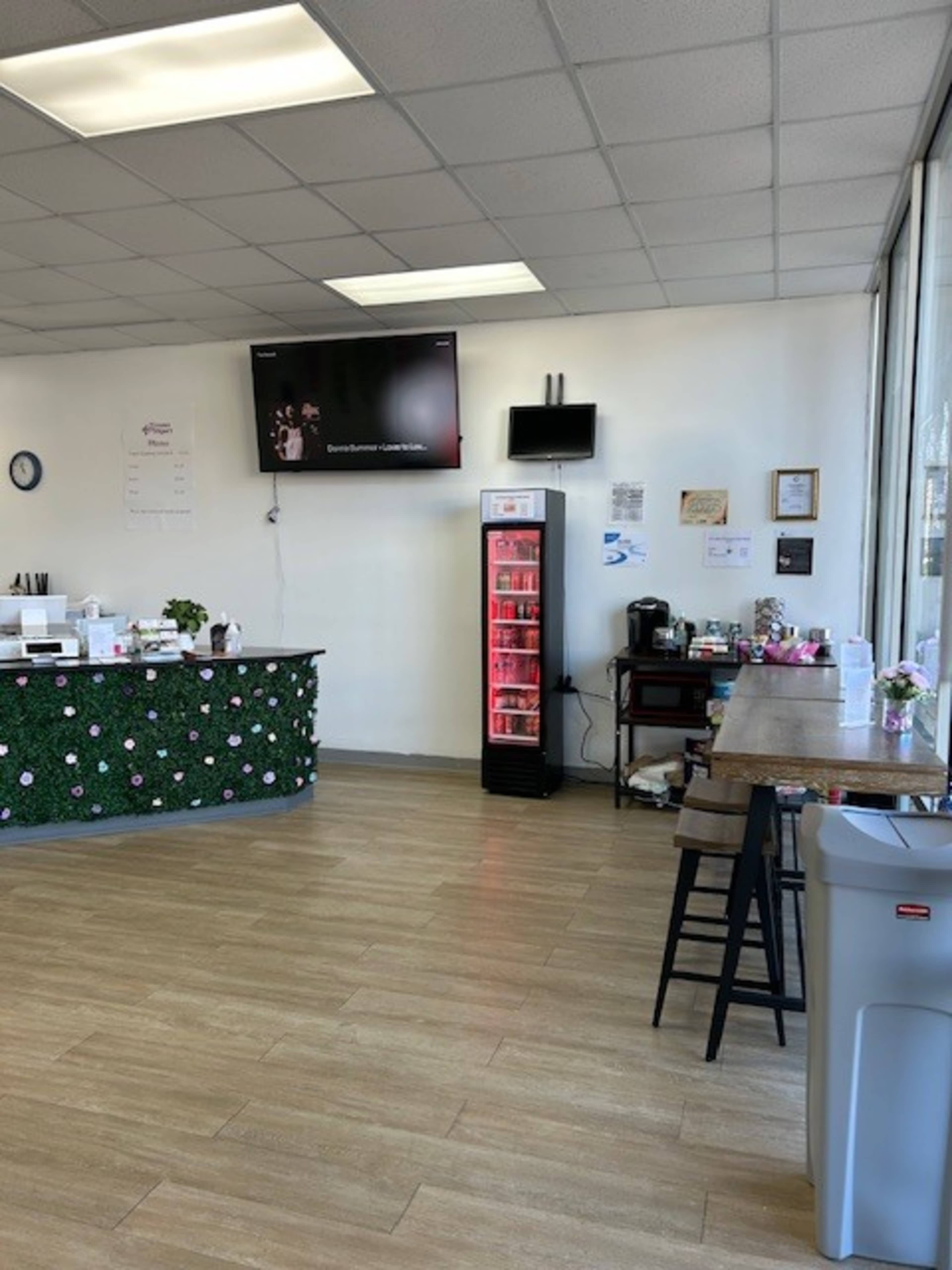 The image shows an interior of a small store or café with a TV on the wall, a colorful counter decorated with greenery, a beverage fridge, and various furniture including a table and a coffee station.